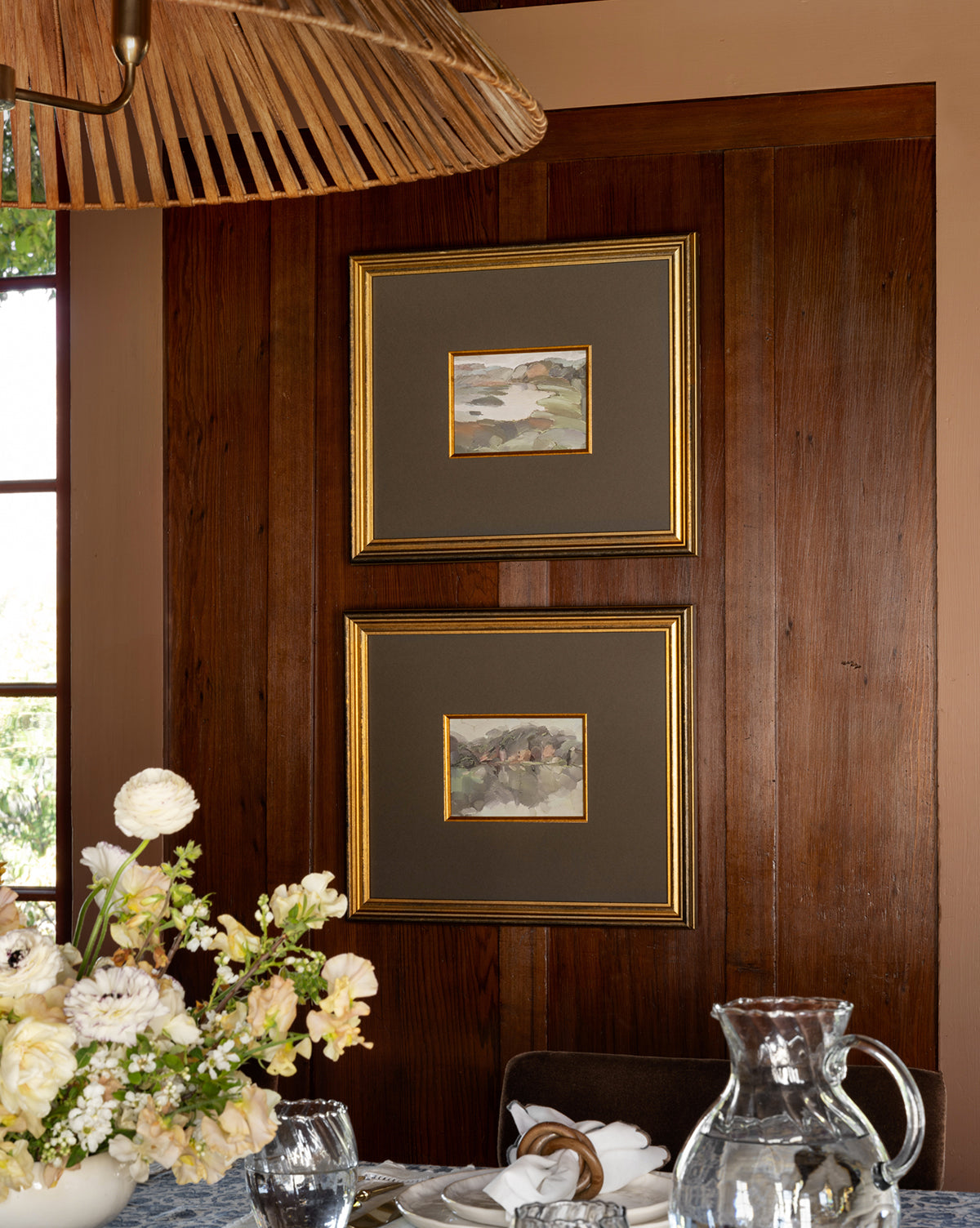 The framed landscape painting Sage Shores II hangs on a wooden wall above a dining table arranged with glassware, white flowers, and napkins; an antique gold frame surrounds the art and a large woven light fixture is visible overhead.
