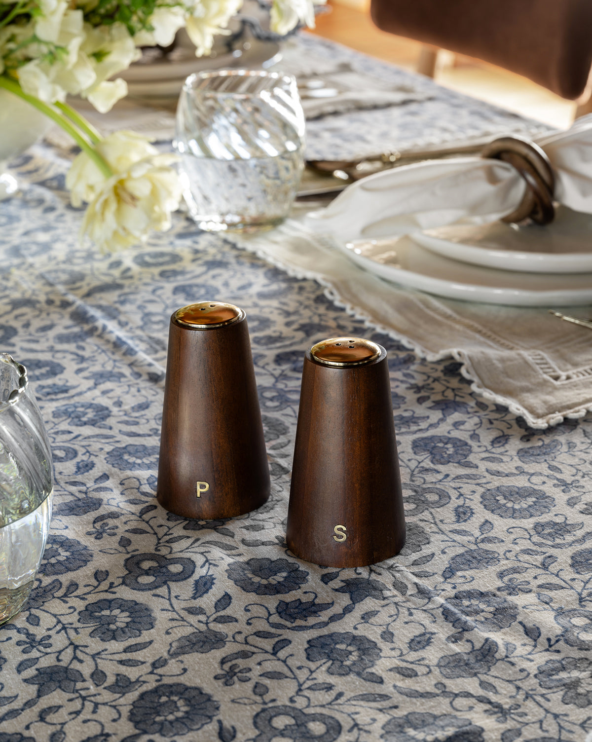 The Brasserie Salt & Pepper Shakers (Set of 2) rest atop a blue floral tablecloth, bringing classic kitchen charm to a set dining table with glassware, plates, napkins, and a bouquet of white flowers.