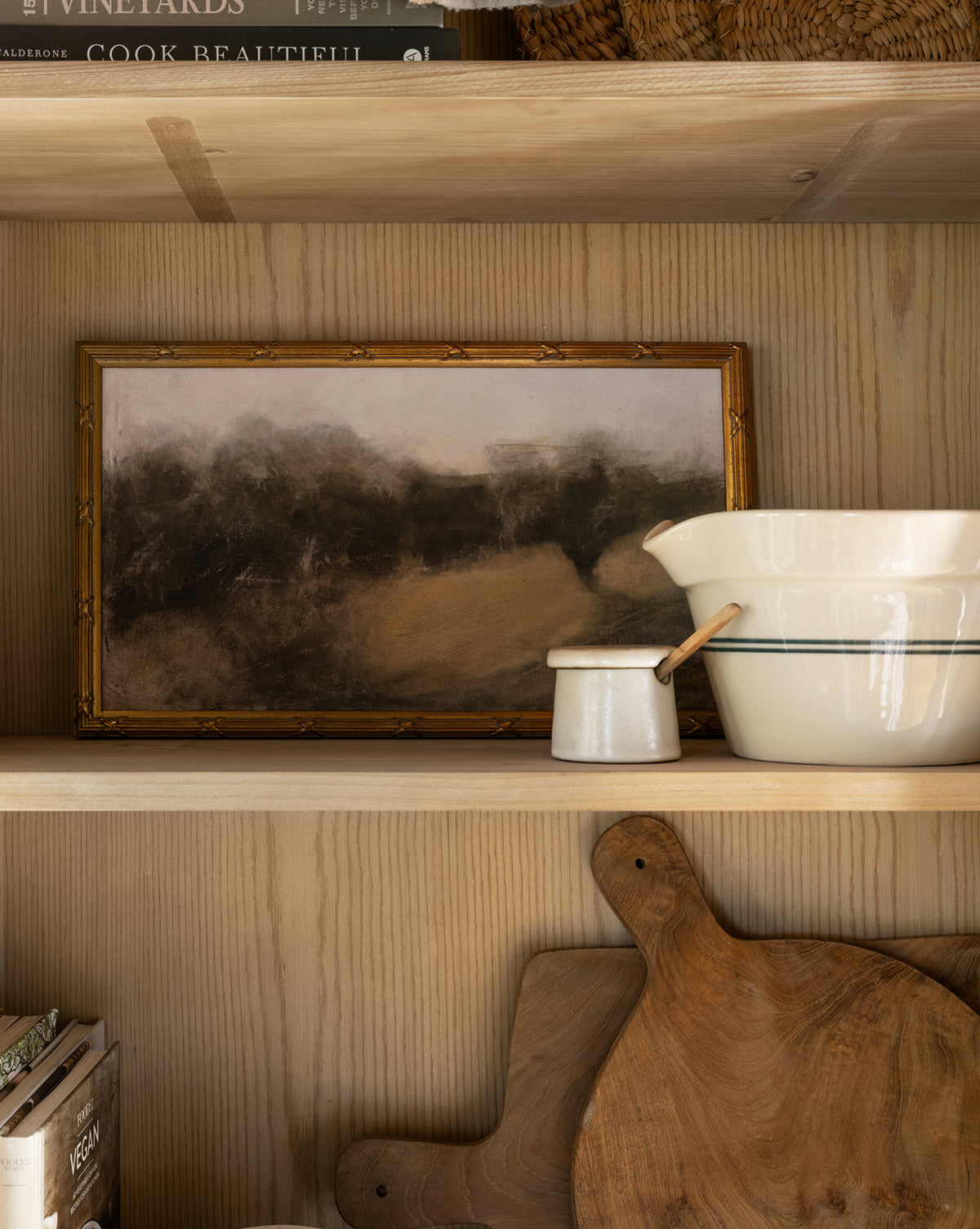 A wooden shelf displays a framed landscape painting, a large white pitcher, a small enamel cup, and two wooden cutting boards. A wooden spoon and the Malco Salt Cellar add charm, with books peeking from the top and bottom shelves.