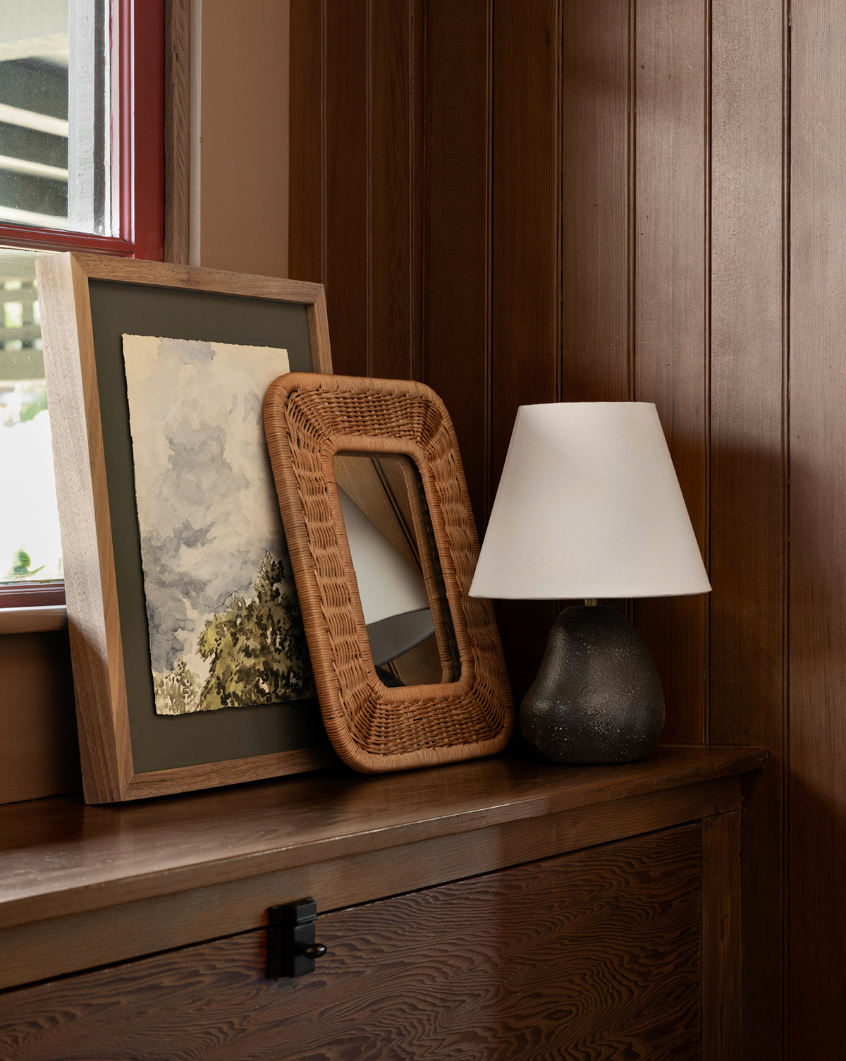 The Head in the Clouds framed art print on paper sits with a wicker mirror and a black table lamp on a dark wooden chest against a wood-paneled wall by the window.
