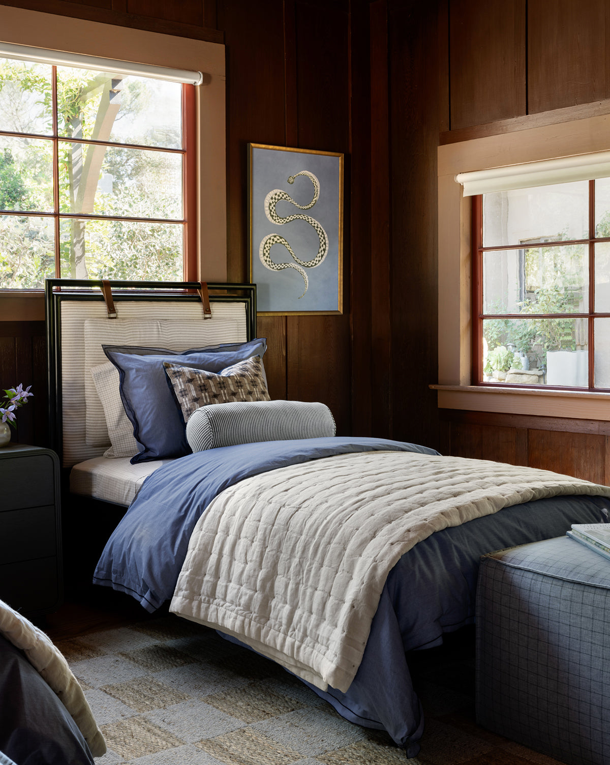 A cozy bedroom with wood-paneled walls, a single bed dressed in the Talcott Quilt with blue and white bedding, large sunlit windows, and a framed snake artwork hanging between them.