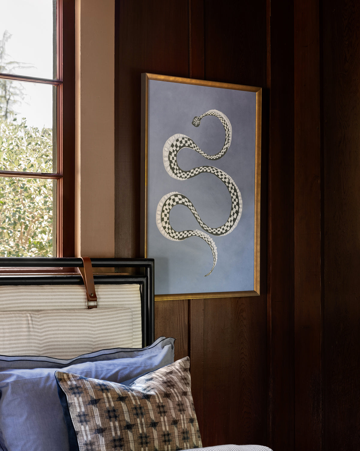 Serpent l, a gold-framed artwork of a white and black patterned snake on a blue background, hangs above a bed with blue and patterned pillows near a window—perfect as unique snake wall art for childrens room decor.