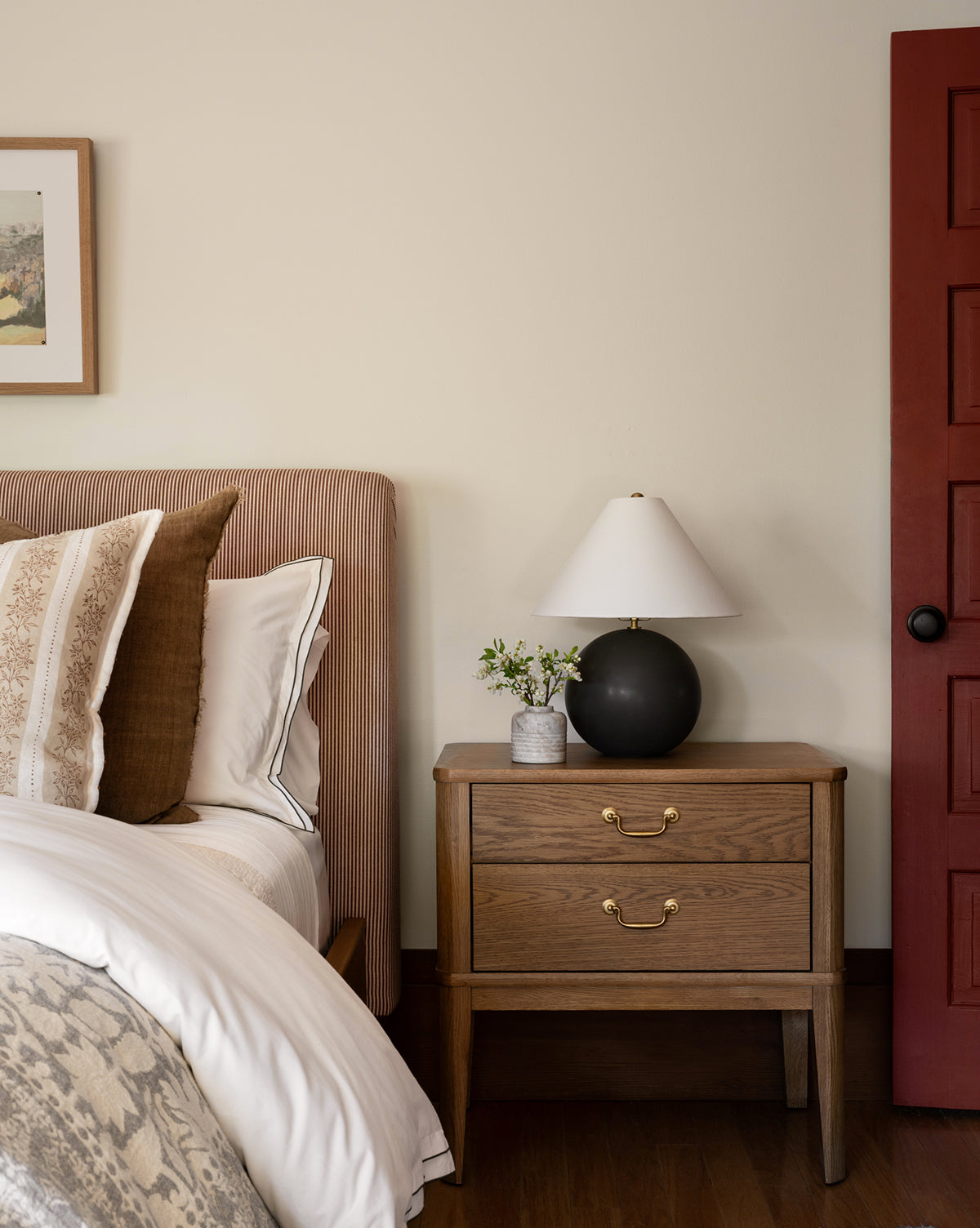 A cozy bedroom displays a bed with striped and patterned pillows, the Alessandra Nightstand with a lamp, a small vase of flowers, and a red door, all set against a neutral wall adorned with framed art, by McGee & Co.