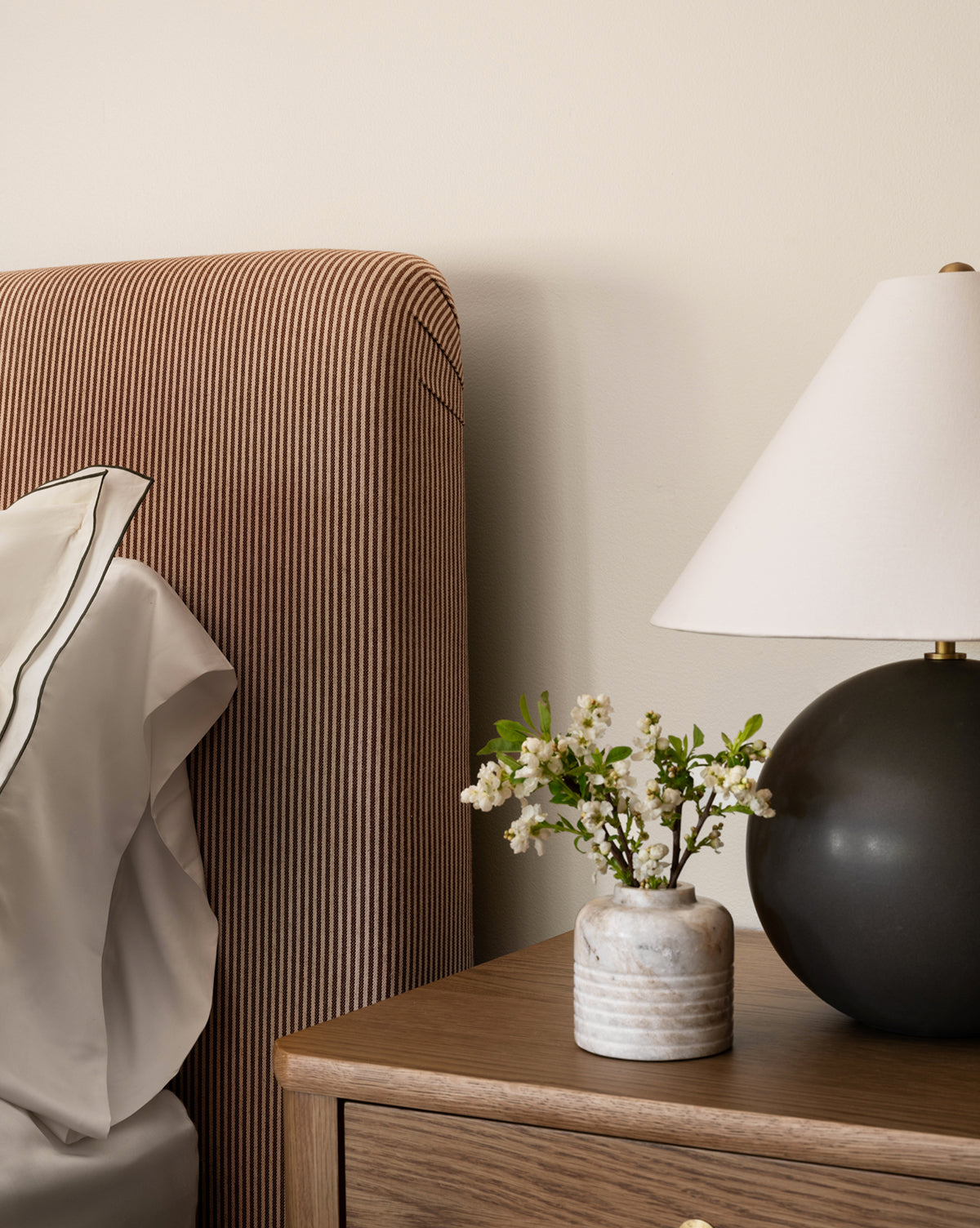 A close-up of a bedside table featuring the Stinson Marble Bud Vase with flowers and a black lamp with a white shade. Part of a bed with a brown striped headboard and white pillow appears on the left, by McGee & Co.
