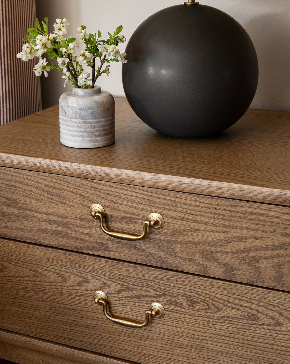 The Alessandra Nightstand in honey oak, featuring brass handles, is topped with a small white-flowered ceramic vase and a round matte black decor piece, creating an elegant display against a neutral wall, by McGee & Co.