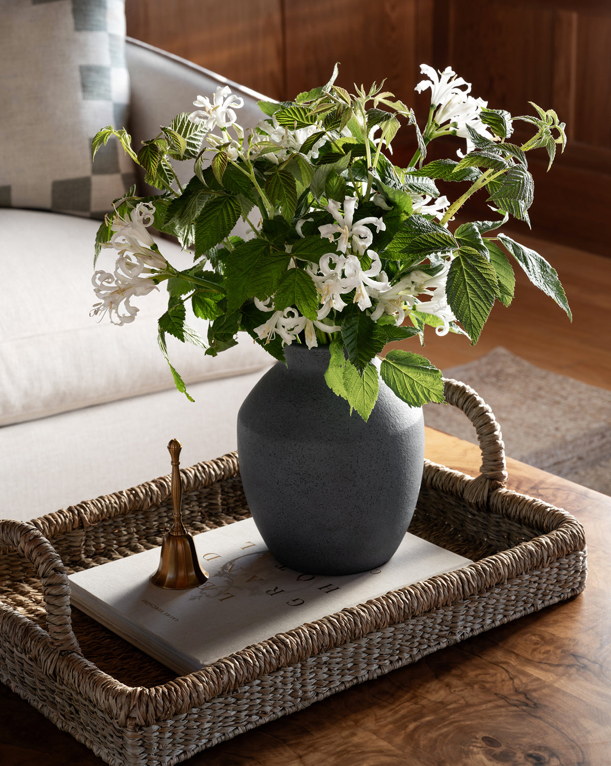 The Liesl Tray, crafted from woven mendong grass, holds a stone vase with white flowers and green leaves atop a book. A small brass bell sits beside it on a wooden table next to a light-colored couch for organic home decor.