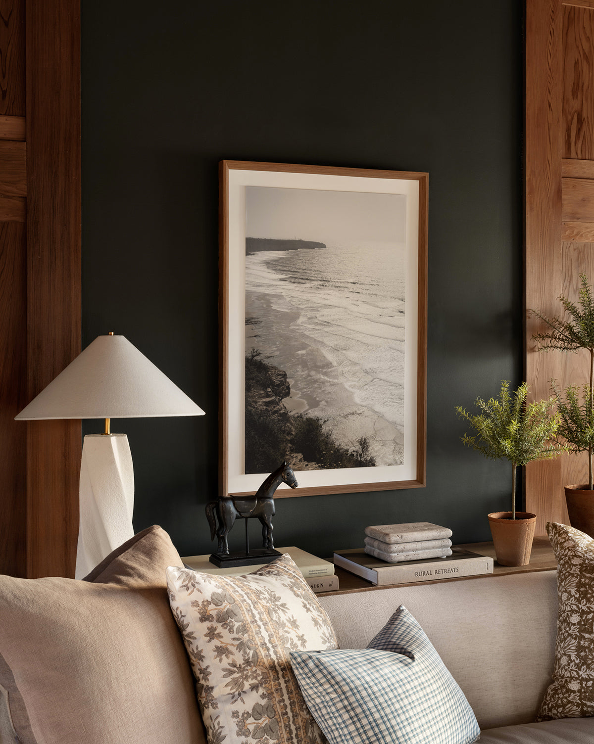 A cozy living room features a beige sofa, patterned pillows, a white lamp, books, a black horse figurine, potted plants, and the striking Shimmer of Wave coastal wall art in a natural wood frame on a dark paneled wall.