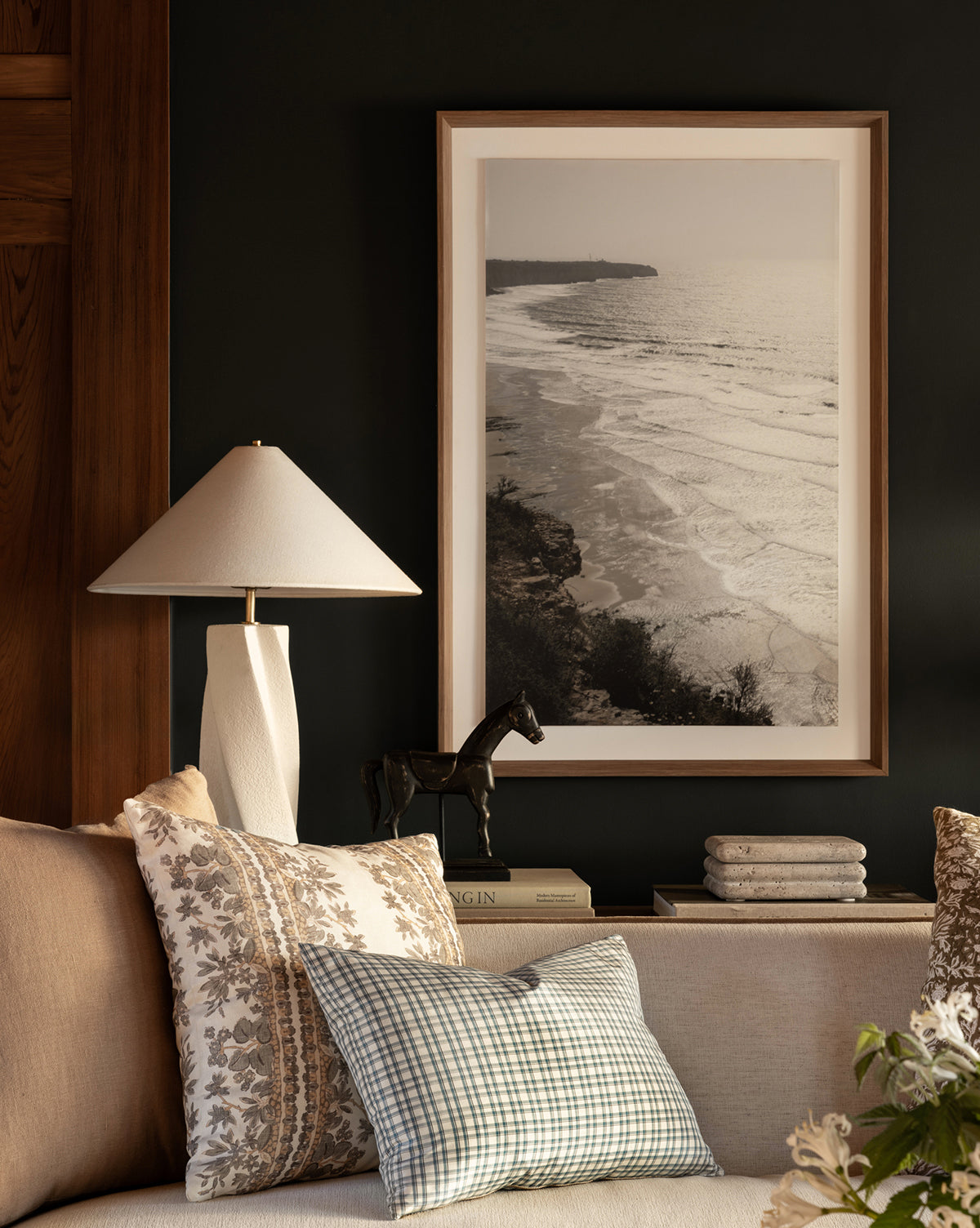 A cozy living room corner features a beige sofa, patterned pillows, stacked books, a horse figurine, and the Bettencourt Table Lamp with a linen shade next to a framed black-and-white coastal photo on a dark wall, by McGee & Co.