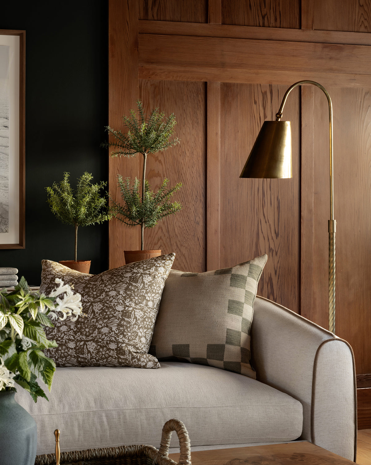 A light gray sofa with two patterned throw pillows sits before a wooden-paneled wall. Two potted plants and the Saylor Floor Lamp offer elegant lighting, creating a cozy, sophisticated corner in the living room by McGee & Co.