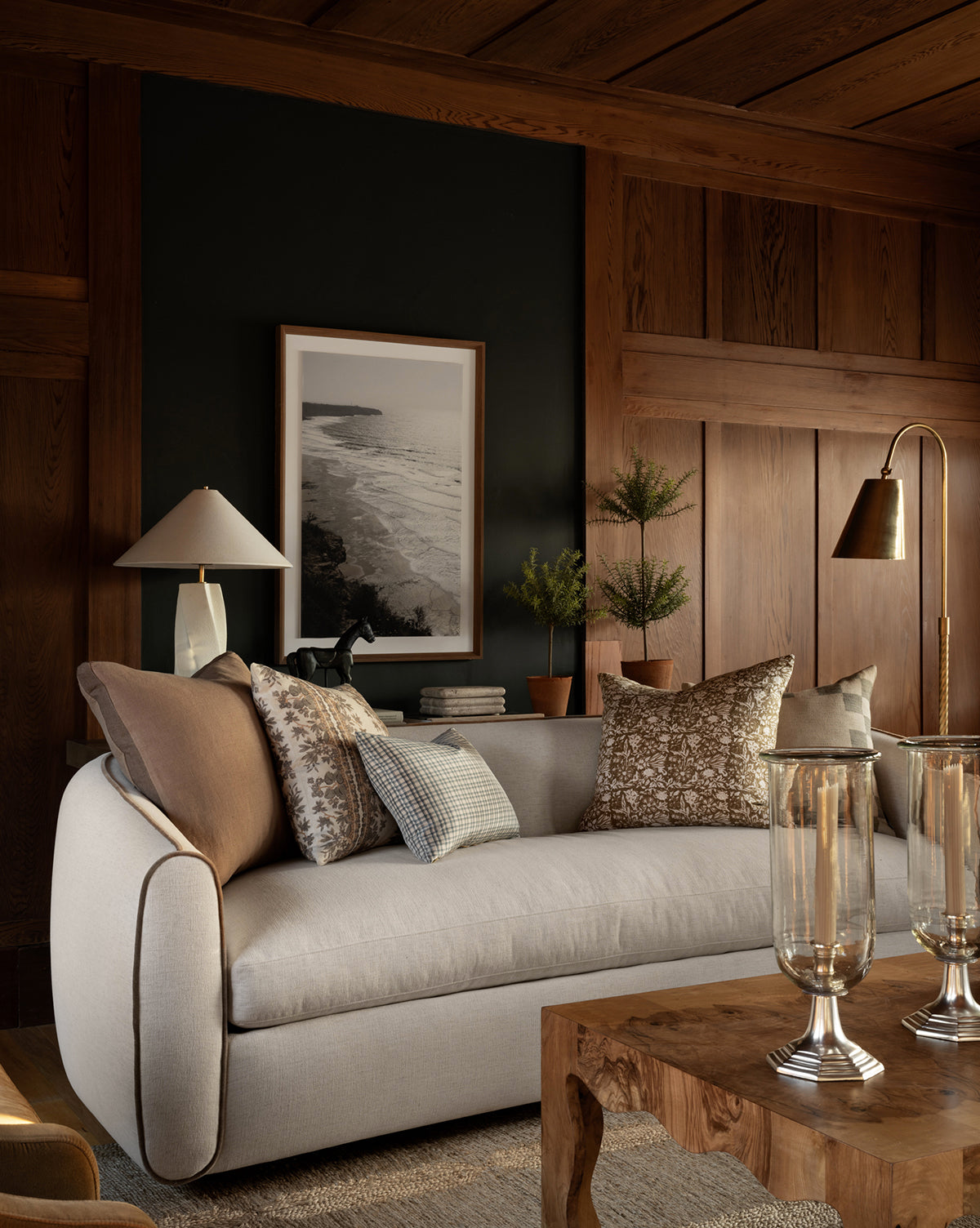 A cozy living room features the customizable Wexler Sofa 90 by Rowe Fine Furniture, Inc., with patterned pillows, a wooden coffee table with glass decor, a table lamp, potted plants, and a framed landscape photo on a dark paneled accent wall—McGee & Co.