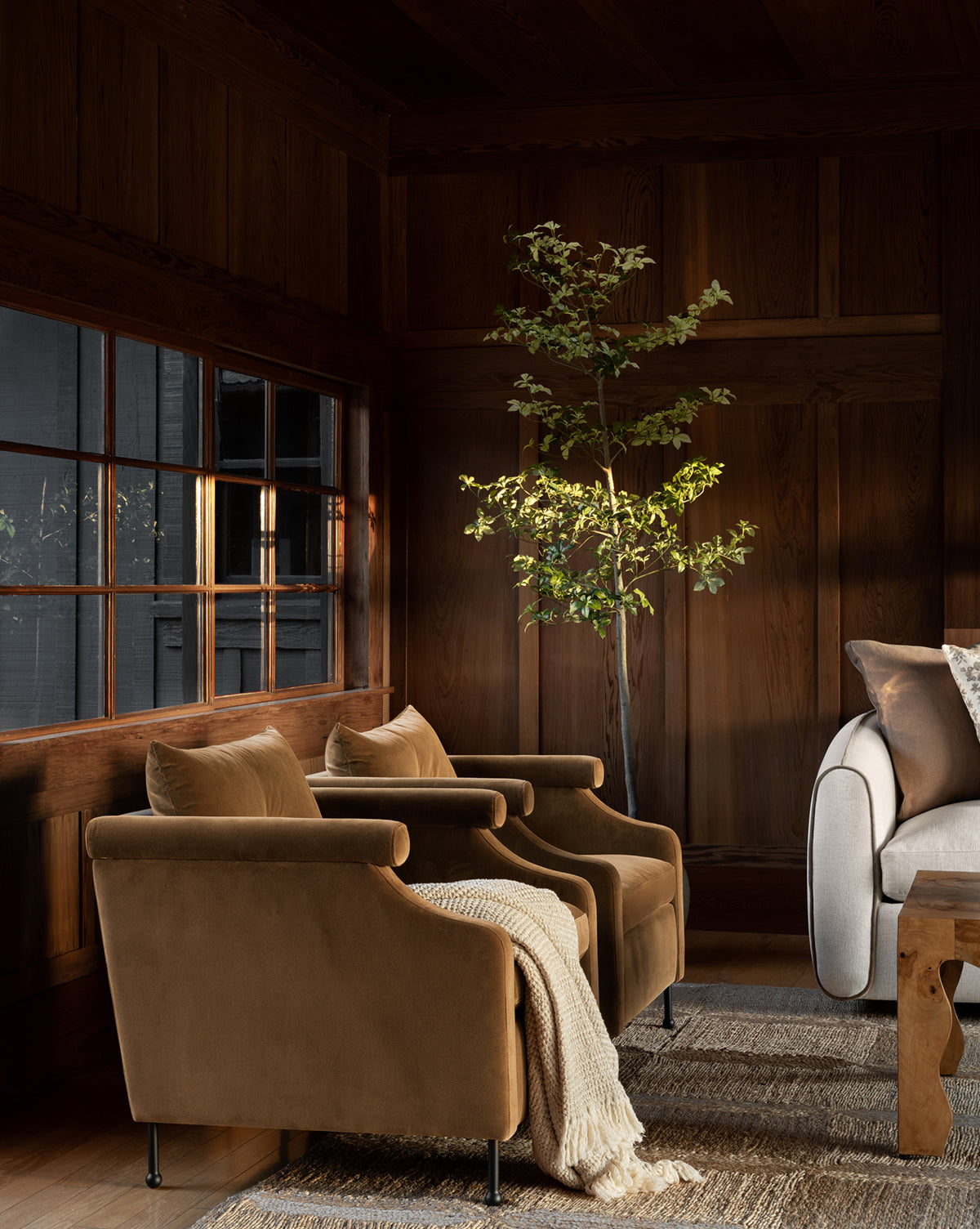 A cozy living room with two Clegg Lounge Chairs featuring iron legs, a light throw blanket, a potted tree, a gray sofa with pillows, wood-paneled walls, and natural light streaming in from large windows by McGee & Co.