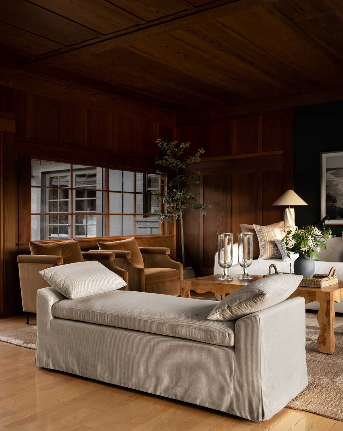 A cozy living room with wooden walls and ceiling features a beige daybed, two brown velvet armchairs, the Scaletta Bench (Ready to Ship) with linen ticking stripe upholstery, a wooden coffee table with candles and flowers, and large windows by McGee & Co.
