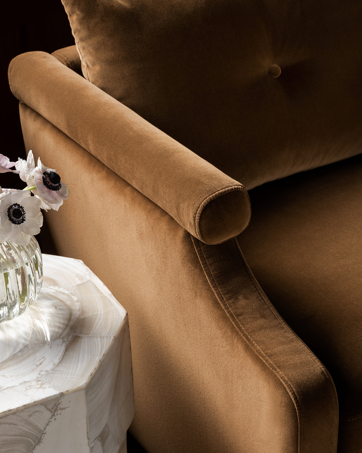A close-up of the Clegg Lounge Chair (Ready to Ship) in brown cotton velvet with a buttoned back cushion, next to a white marble side table displaying a glass vase filled with white flowers by McGee & Co.