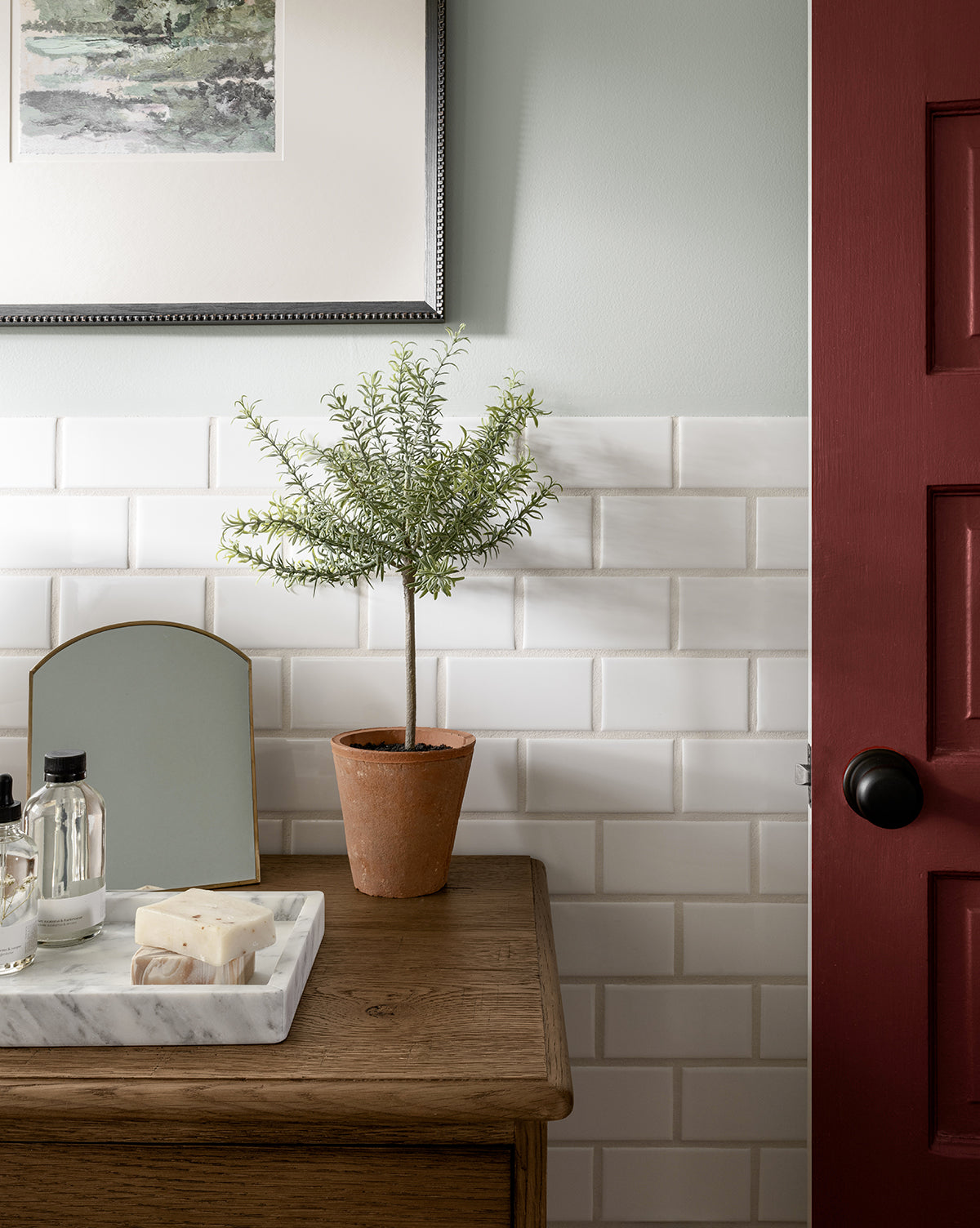 A Potted Faux Rosemary Topiary in a terracotta pot rests on a wooden table with toiletries atop a marble tray, against a white subway tile wall. A red door with a black knob and framed art above add to the cozy ambiance.