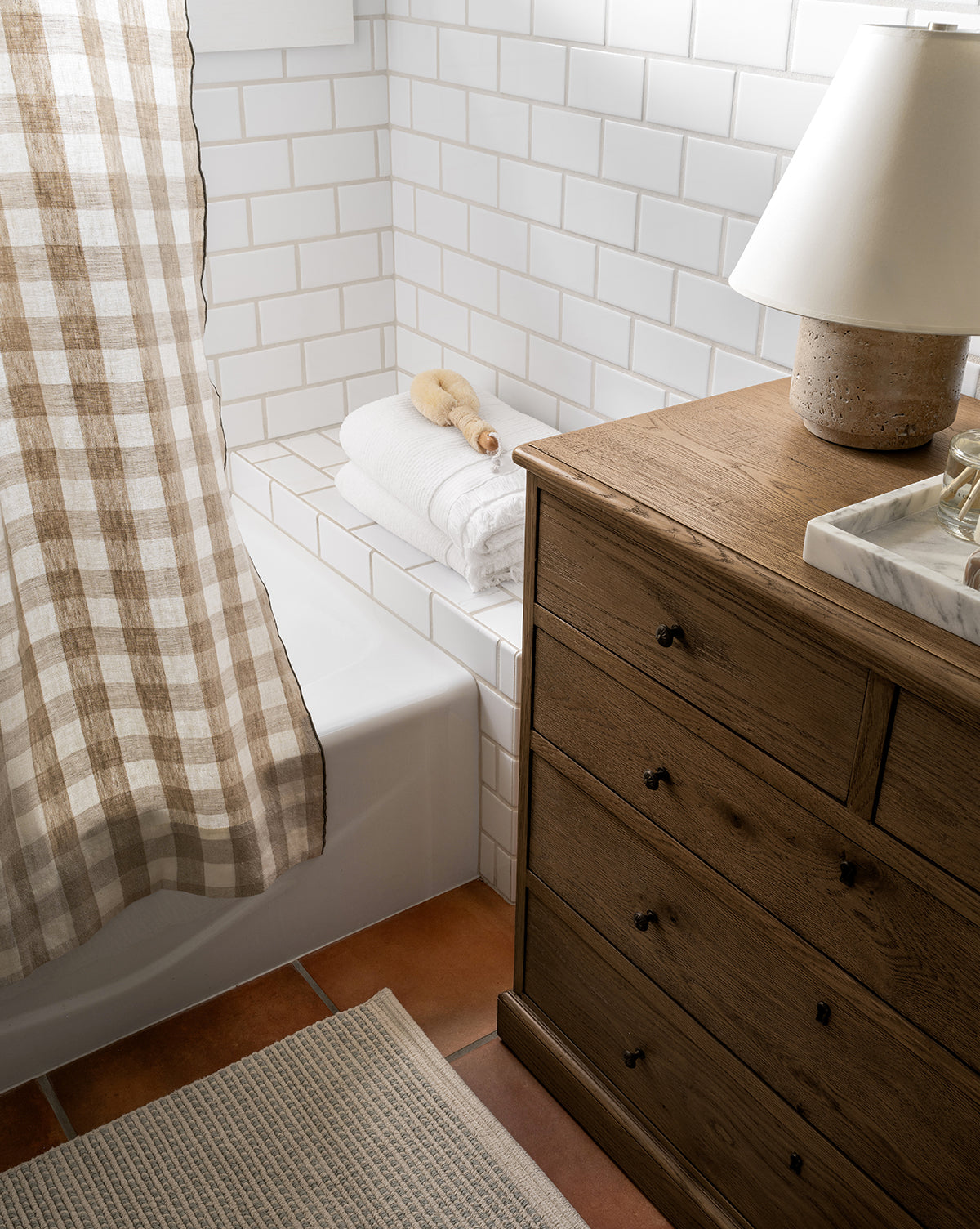 A bathroom with white subway tile, a tub partially covered by a brown and white checkered shower curtain, 100% cotton towels and bath brush on the tub edge, an Emina Bath Mat, a wooden dresser, and a lamp on a marble tray.