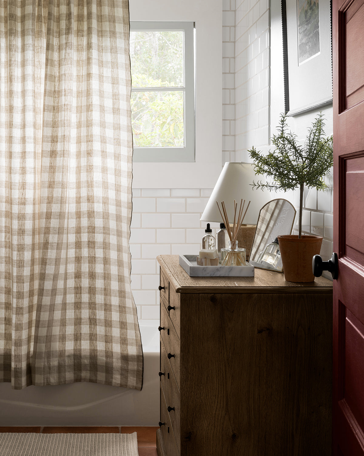 A bathroom with white subway tiles, a wooden vanity, and potted plant. Sunlight streams in as the Pennie Shower Curtain in beige checkered print hangs by the tub, complementing stylish decor on the countertop.