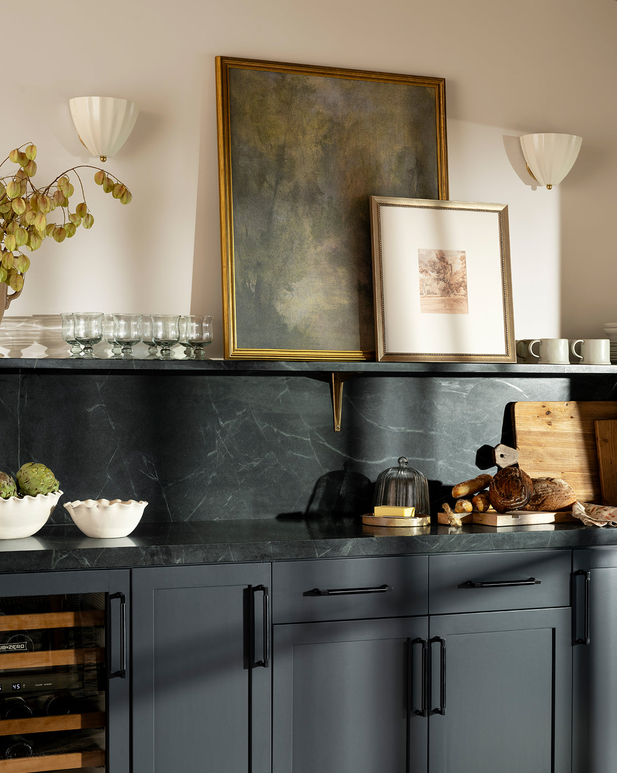 Modern kitchen featuring Dream State artwork, dark cabinets, black marble surfaces, wooden cutting boards, ceramic bowls, glassware, two wall sconces, and dried foliage for a stylish finish, by McGee & Co.