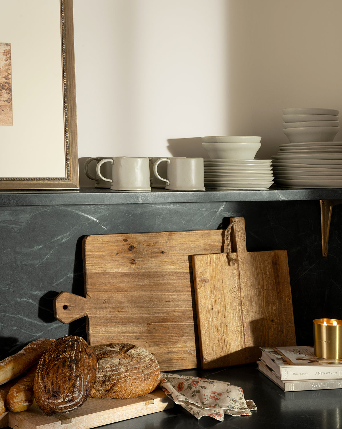 A kitchen counter displays rustic bread, wooden cutting boards, a floral napkin, stacked white dishes, the Aiden Soft Gray Bowl, mugs, a framed picture, books in a stack, and a lit candle set on a dark stone surface, by McGee & Co.