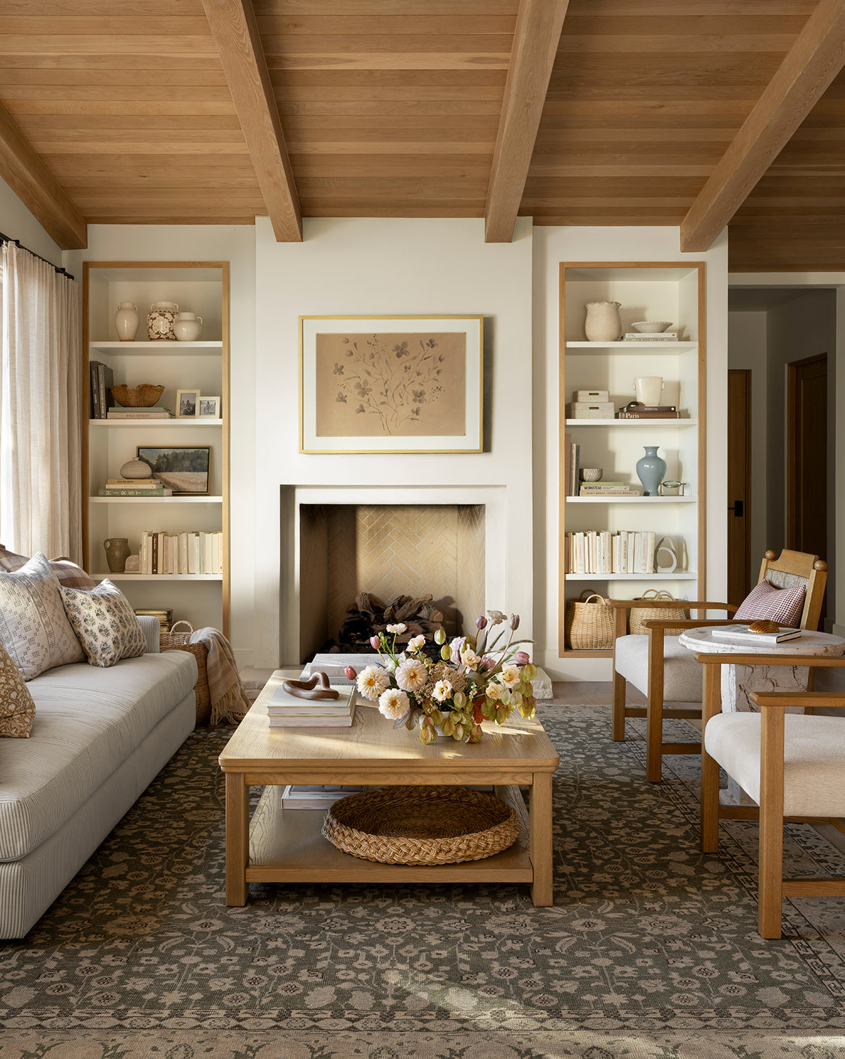 A cozy living room with wooden ceiling beams, built-in shelves, a framed artwork above the fireplace, a beige sofa, two armchairs, a wooden coffee table with flowers, and the Fernwood Hand-Knotted Wool Rug, by McGee & Co.