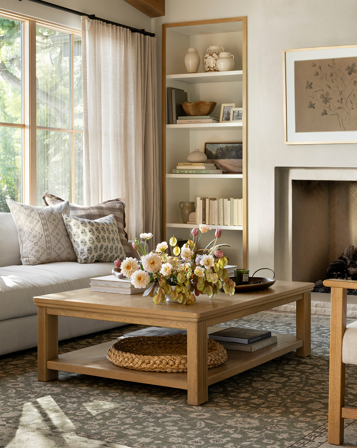 A cozy living room with a light sofa, patterned pillows, an Ambrose Coffee Table by YANG CHENG WOODEN INDUSTRIES INTERNATIONAL (VIETNAM) CO LTD topped with flowers, a woven tray beneath, built-in shelves, fireplace, and large window with sheer curtains. McGee & Co.