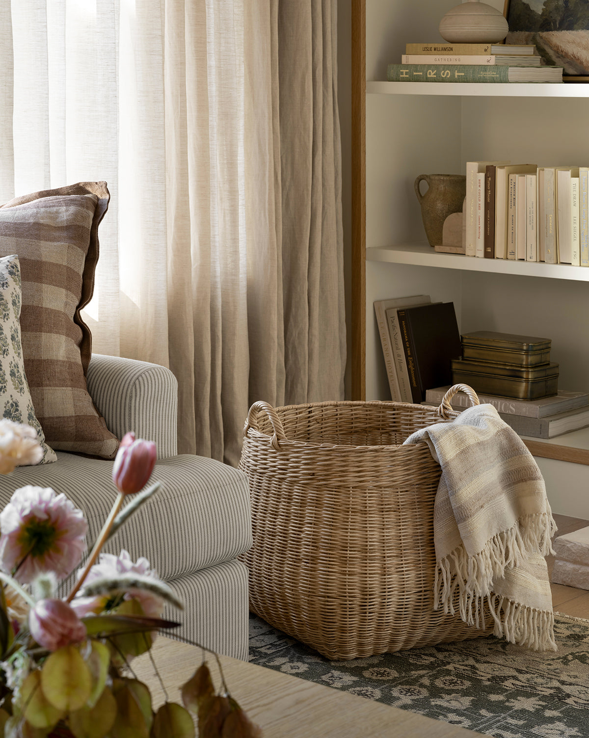 A cozy living room corner with a striped armchair, a plaid pillow, the Boyce Woven Basket holding a fringed blanket, books on white shelves, sheer curtains filtering soft natural light, and pink flowers in the foreground, by McGee & Co.