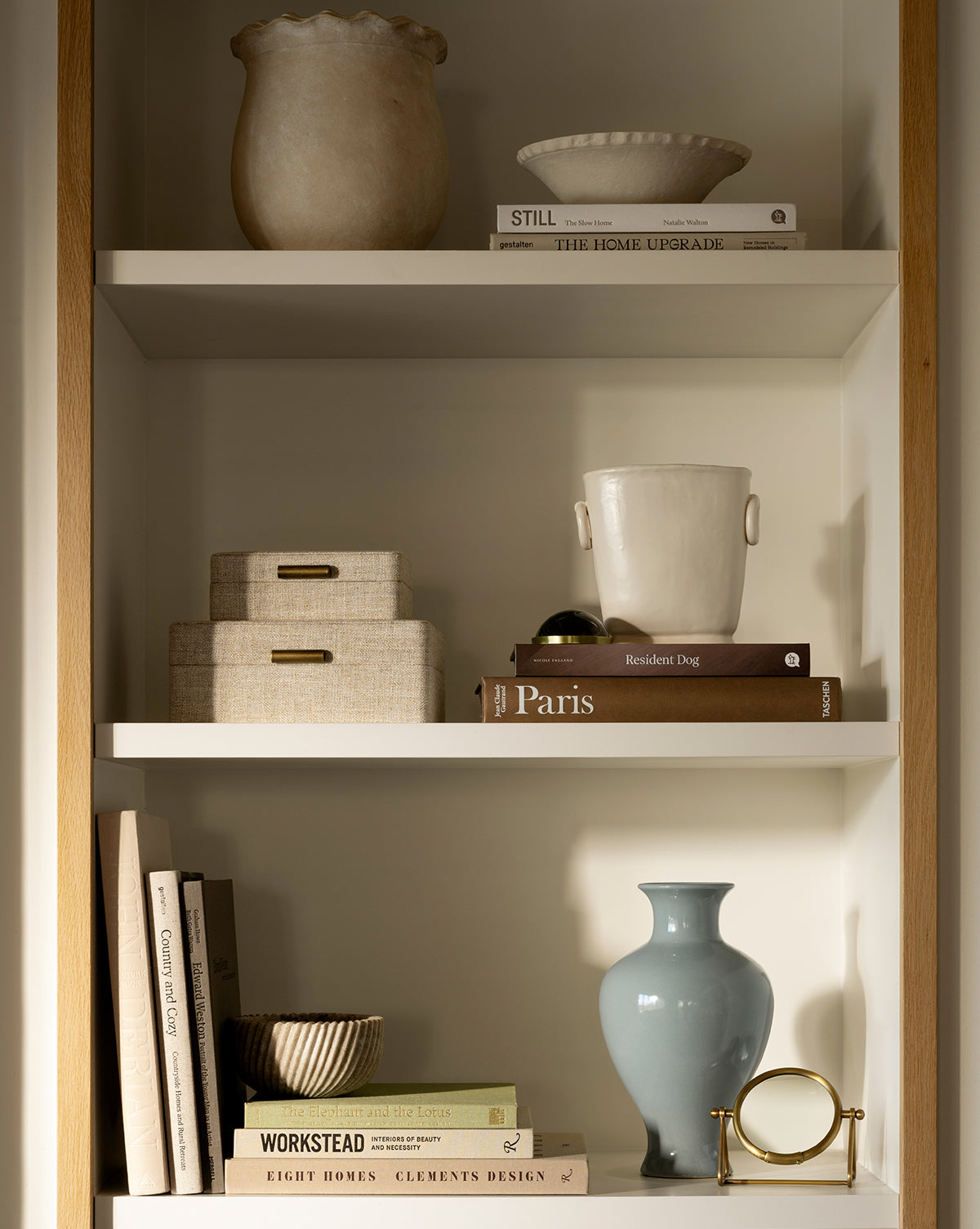 A neatly arranged bookshelf features Swansea Indoor Pot, ceramic vases, stacked books, decorative boxes, and small sculptural objects on three shelves in a warmly lit setting, by McGee & Co.