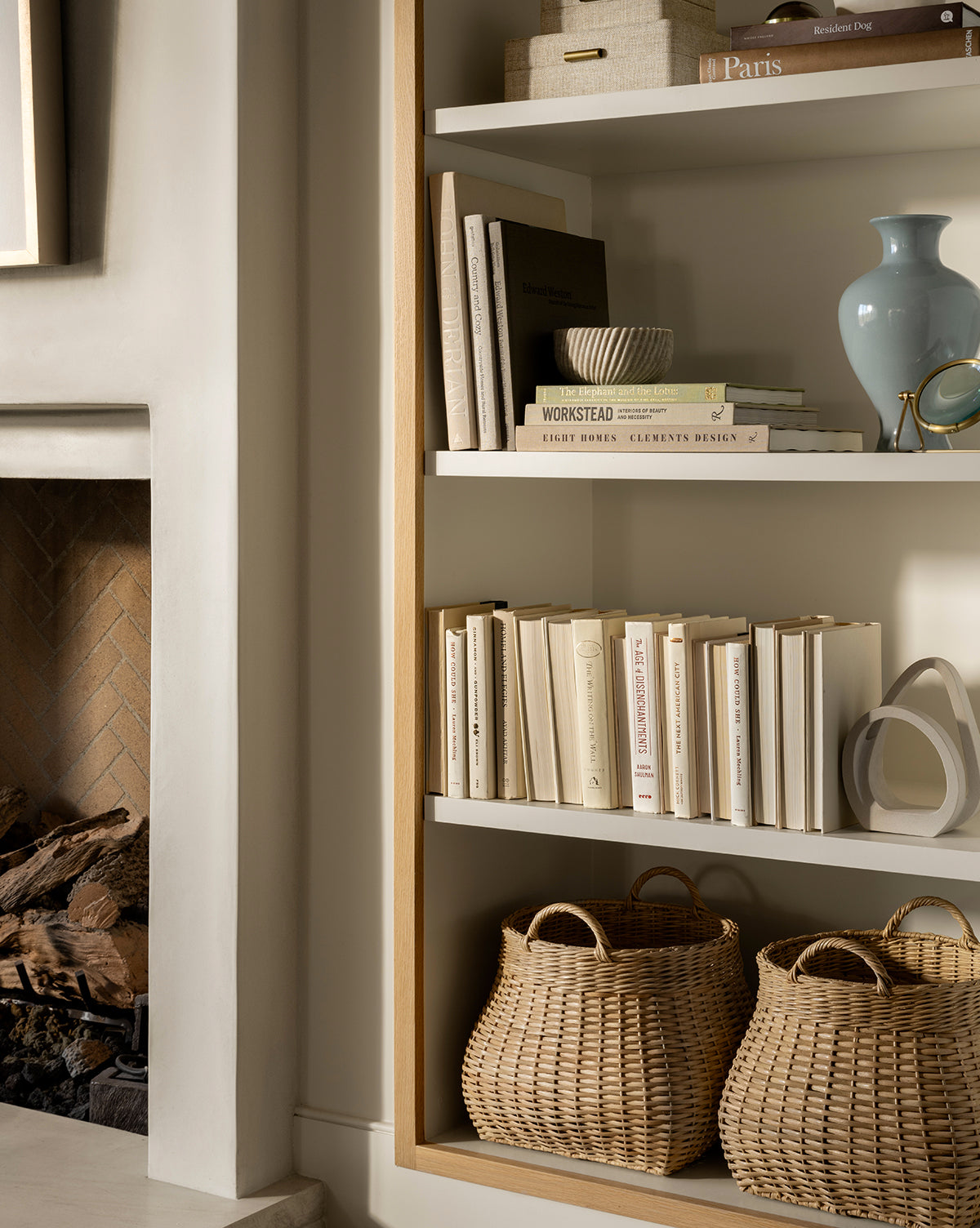 A modern, neutral bookshelf displays stacked books, a blue vase, décor pieces, and two Boyce Woven Baskets on the lower shelf beside a fireplace with stacked logs, by McGee & Co.