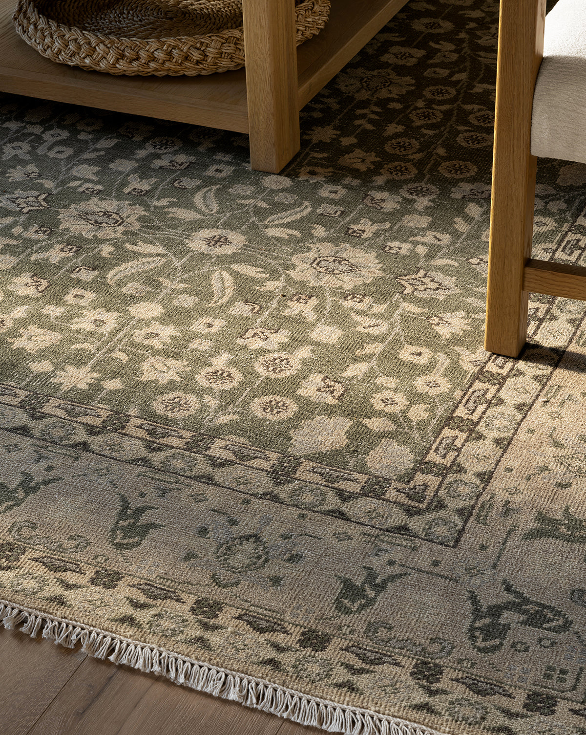 A close-up of the Fernwood Hand-Knotted Wool Rug, showcasing floral patterns in muted green and beige. The rug is accented by a wooden table with a woven basket beneath and a chair, all bathed in natural light, by McGee & Co.