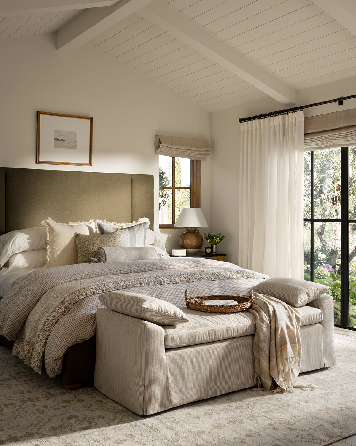 A cozy, neutral-toned bedroom features a large bed with layered bedding and the Scaletta Bench (Ready to Ship) at the foot, topped with a throw and tray, plus a nightstand, lamp, wall art, and big windows with sheer curtains overlooking greenery by McGee & Co.