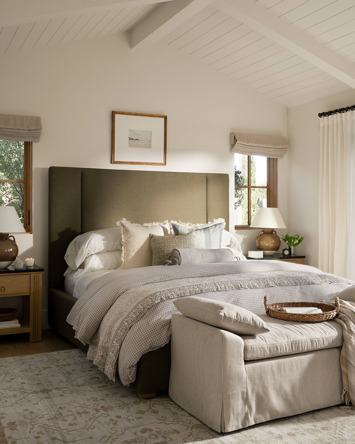 A cozy bedroom features a plush bed with neutral bedding, pillows, and a soft throw. The Lexington Hand-Knotted Wool Rug anchors the space, paired with matching nightstands, lamps, art above the bed, and plenty of natural light, by McGee & Co.