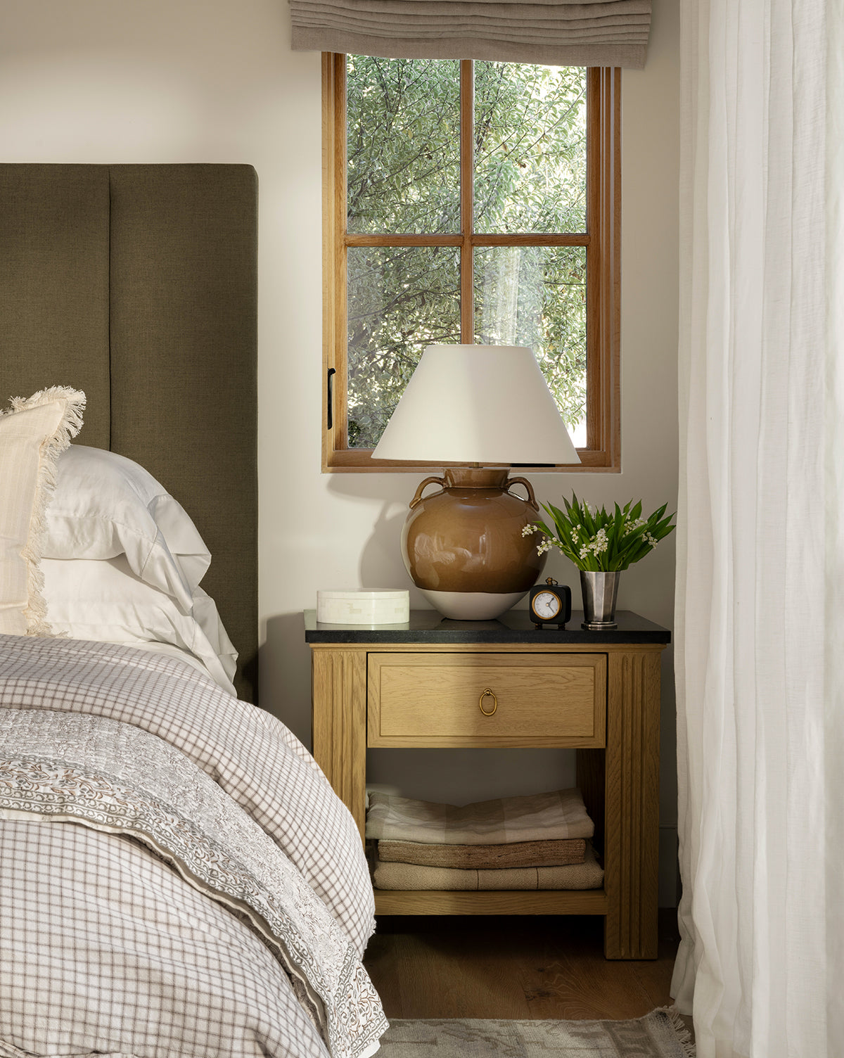 A cozy bedroom scene shows a neatly made bed and an Ambrose Nightstand in Drift Oak from YANG CHENG WOODEN INDUSTRIES INTERNATIONAL (VIETNAM) CO LTD, topped with a lamp, brown ceramic vase, alarm clock, and plant as sunlight streams through the window above—McGee & Co.
