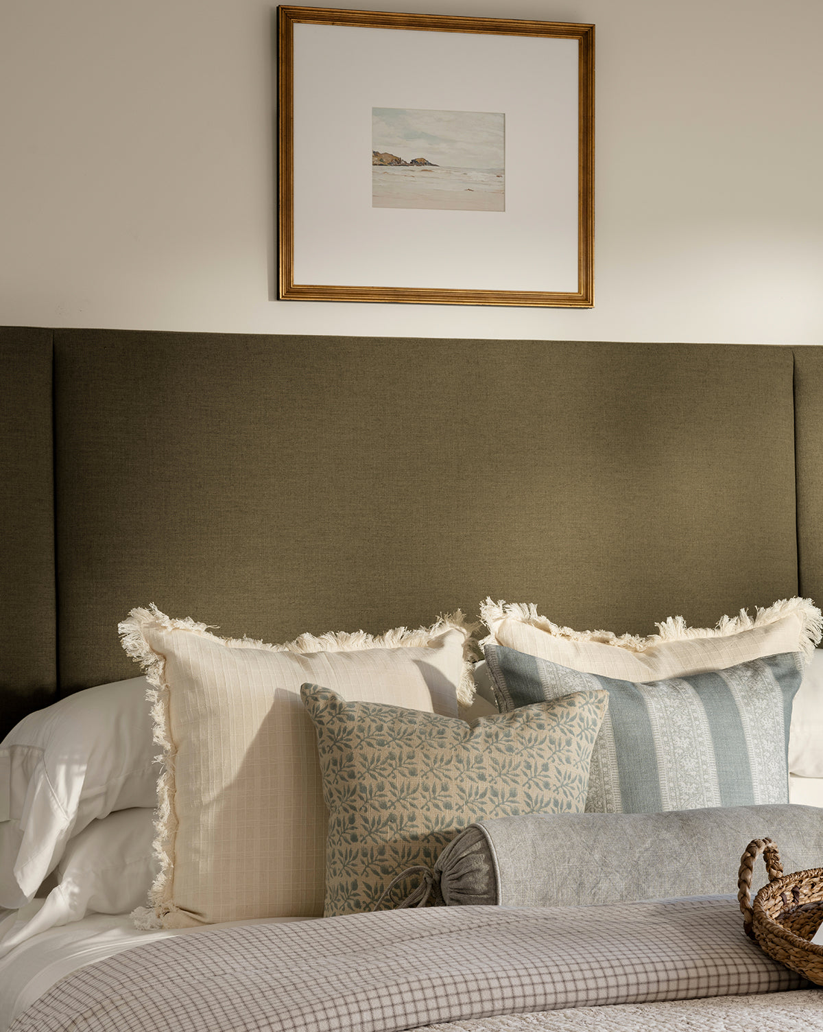 A neatly made bed with layered neutral-toned pillows sits against an olive green upholstered headboard. Above the bed, a framed giclee print titled Shoreline Rocks hangs on a light-colored wall by McGee & Co.