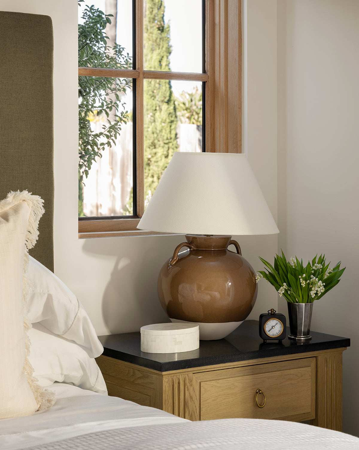 A cozy bedroom scene with a wooden nightstand holding a brown lamp, the Kya Clock, a round marble box, and a vase of white flowers beside the bed, with greenery visible through the window by McGee & Co.