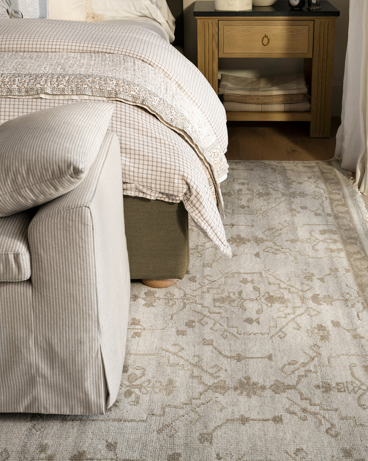 A cozy bedroom corner with a striped armchair, a neatly made bed with plaid and lace bedding, folded blankets on the nightstand shelf, and the Lexington Hand-Knotted Wool Rug crafted by artisans, by McGee & Co.