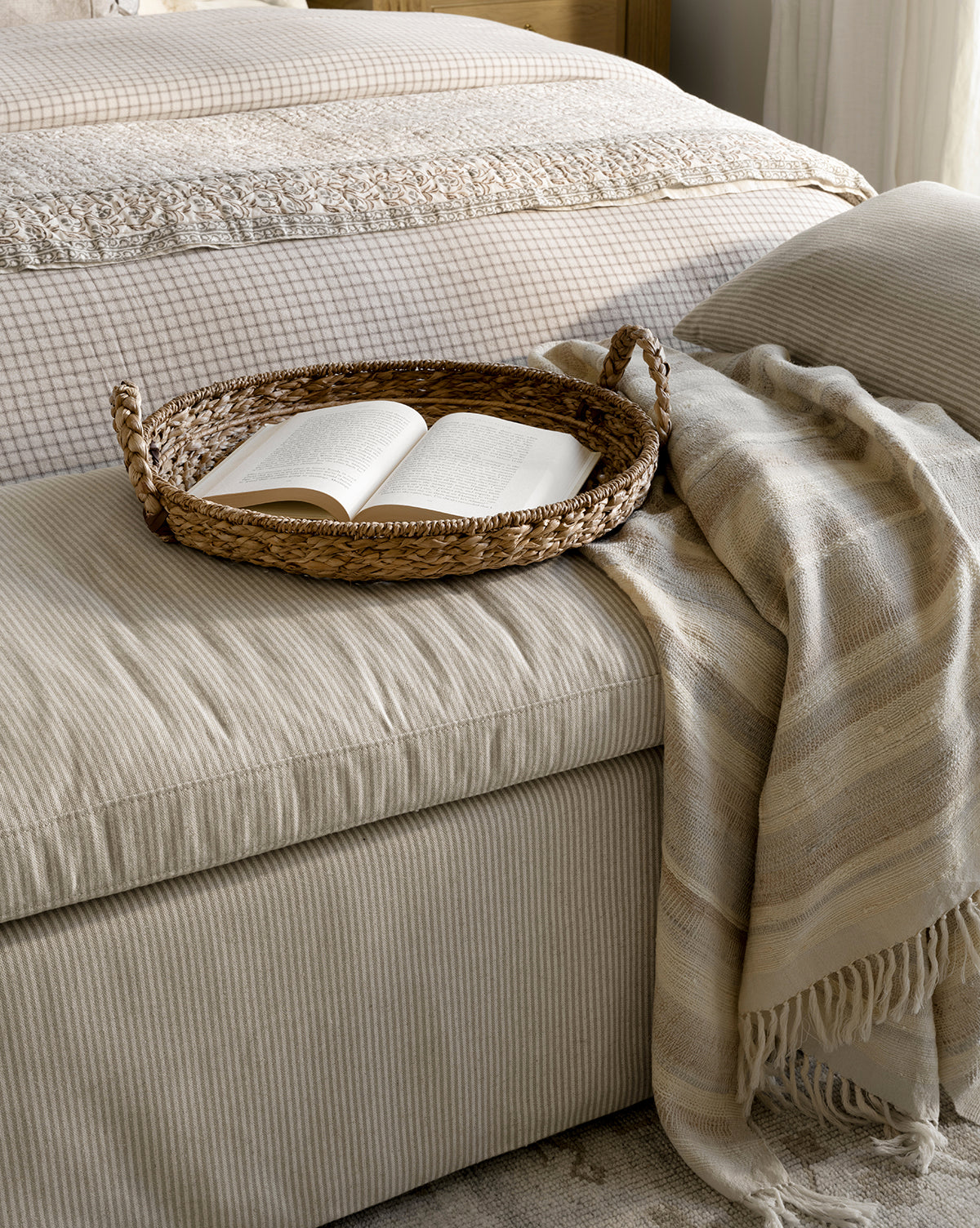 A woven tray with an open book sits on a beige striped ottoman topped with the Laney Stripe Throw by TAL HANDWOVEN CLASSICS LLP, beside a bed dressed in a checked duvet and lace-trimmed linens, McGee & Co.