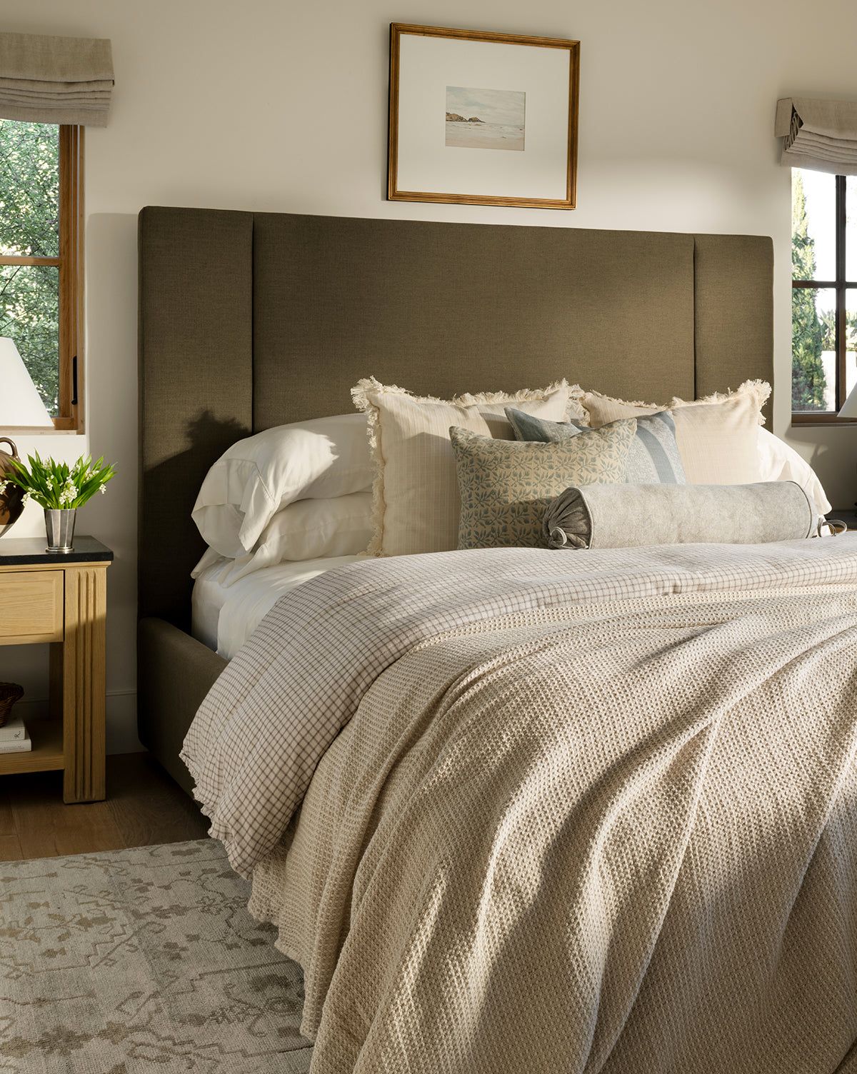A neatly made bed with layered neutral bedding, decorative pillows, a large padded headboard, and the Penedo Duffy Oversized Throw creates a cozy look. Nightstands with lamps and flowers, soft window light, and a framed picture above complete the scene—McGee & Co.