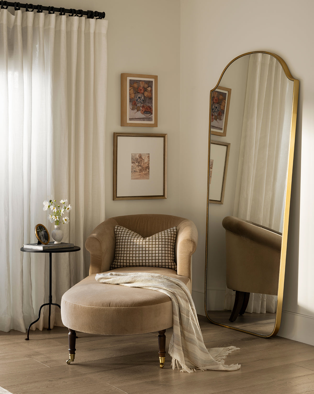 A cozy corner with a beige velvet chair, checkered pillow, striped throw, and a round side table with flowers sits by framed art and the Sussex Floor Mirror, by McGee & Co. Light curtains drape the window.
