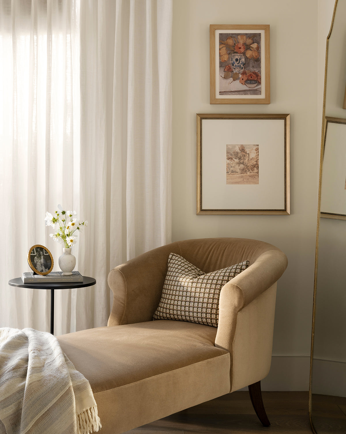 A cozy reading nook features a beige chaise lounge, patterned pillow, striped blanket, round side table with flowers and photo, white curtains, and the Harewood Park print in a distressed silver frame on the cream wall, by McGee & Co.