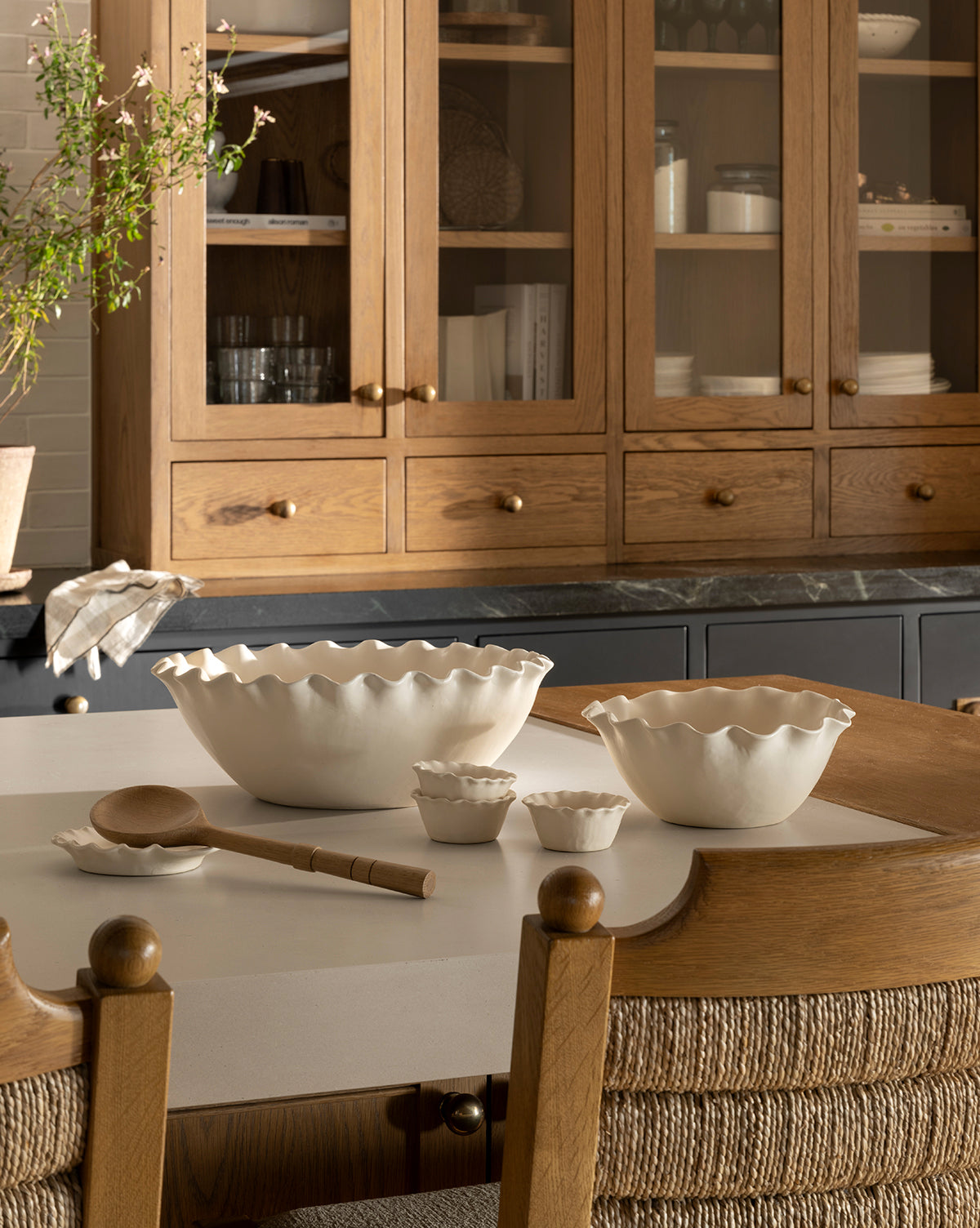 A kitchen with wooden cabinets, glass doors, and a marble countertop features a white island displaying nested Olindo Serving Bowls (Set of 2) with scalloped edges, matching cups, a wooden spoon, and two woven-back chairs by McGee & Co.