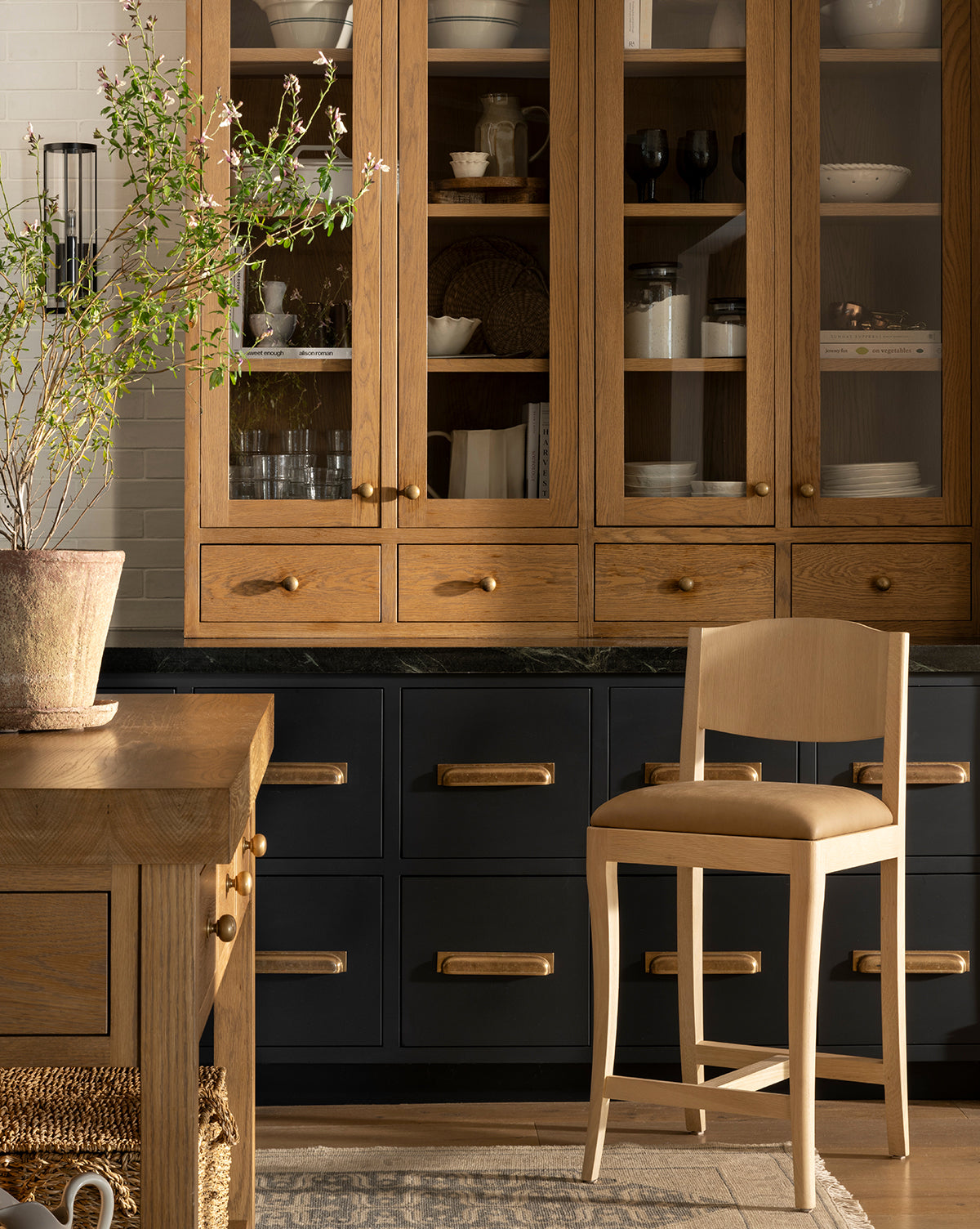A kitchen features wooden cabinets, glass-fronted uppers, black drawers with brass handles, a Makerspalm Shepherd Counter Stool by the white oak island, a wooden table, a potted plant, and soft natural lighting—McGee & Co.