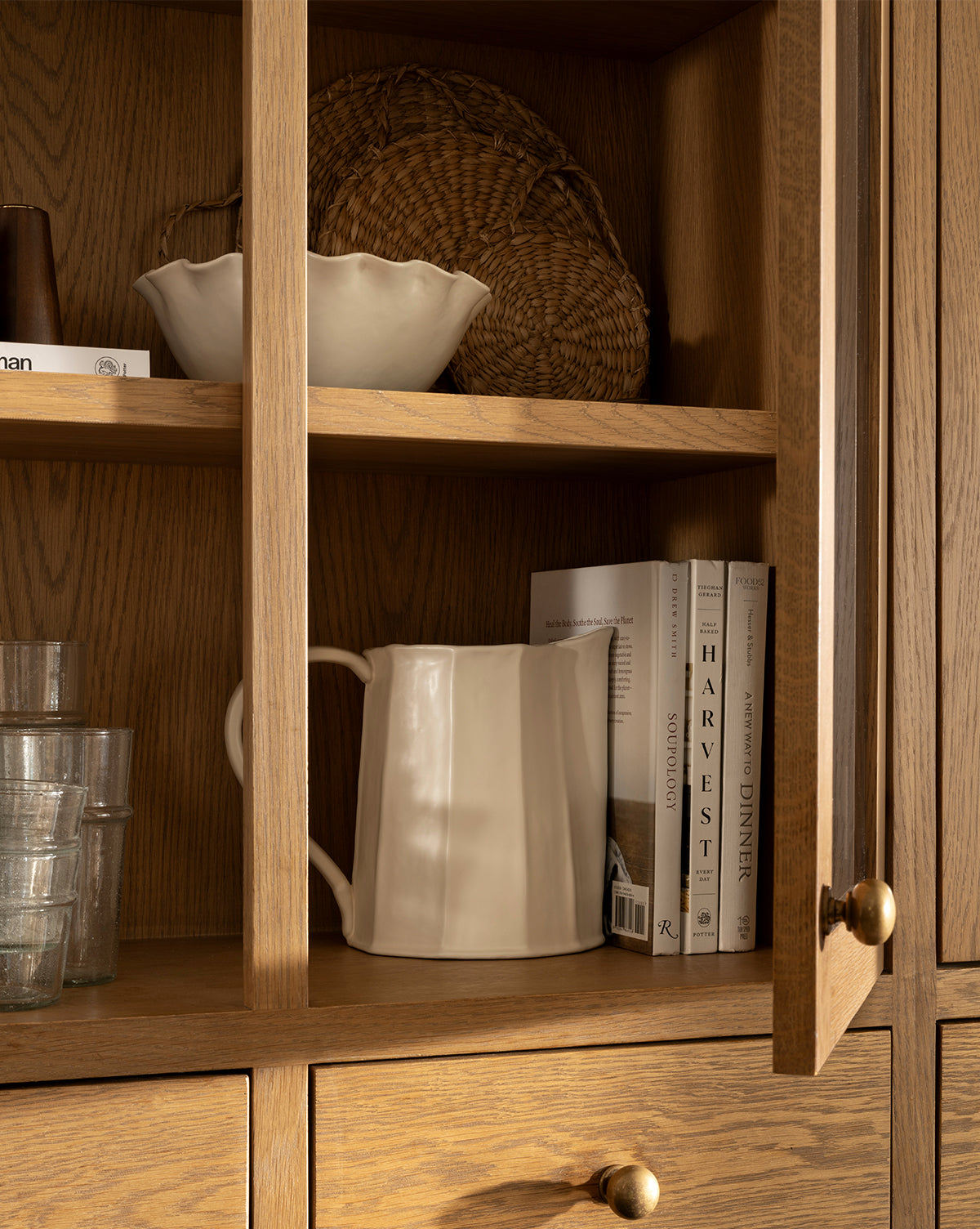 A wooden cabinet with glassware, three books, a white bowl, a woven basket, and the Fielding Ceramic Pitcher displayed on its shelves brings rustic kitchen charm, by McGee & Co. The cabinet door is partially open.