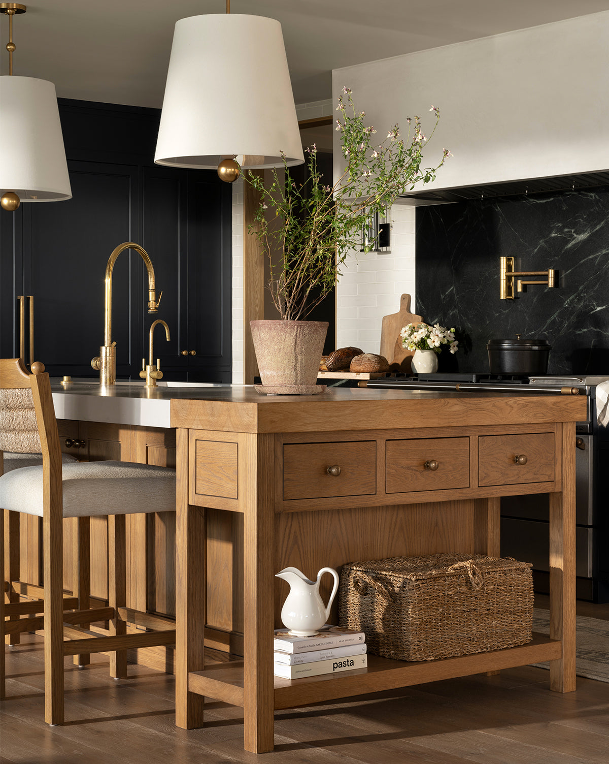 A modern kitchen features a wooden island, gold fixtures, two pendant lights, black cabinetry, a potted plant, wicker basket, and the Lumen Pitcher in white finish on the island shelf near the pasta jar. Marble backsplash and stove in background, by McGee & Co.