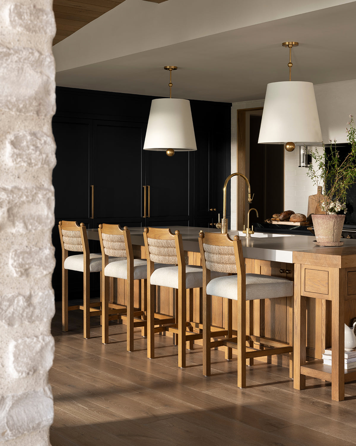 A modern kitchen features a honey oak island, four Makerspalm Alberta Counter Stools with cushions, gold fixtures, black cabinets, two large white pendant lights above the counter, and a potted plant on the island. McGee & Co.