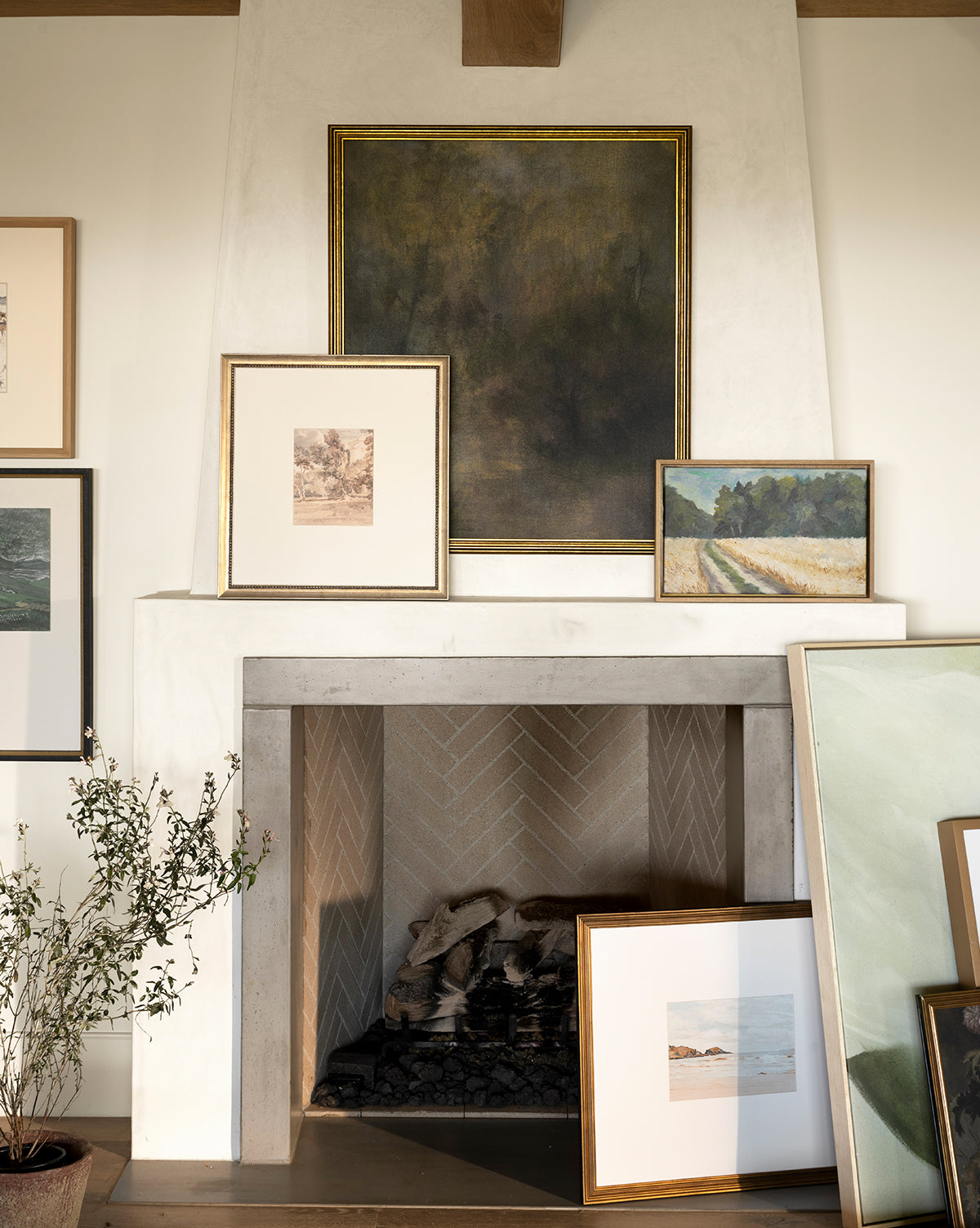 The Harewood Park light-colored fireplace with a herringbone pattern features framed landscapes and abstract art in distressed silver wood frames on the mantel and hearth, with a potted plant on the left, by McGee & Co.