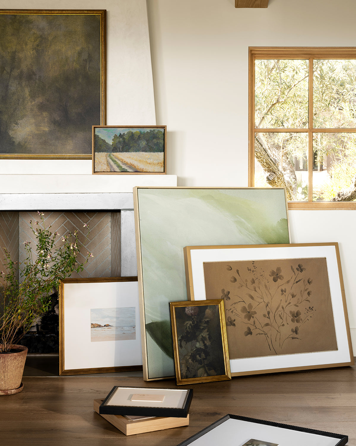 ‘Consider the Wildflowers’ by Katie Mulder, a giclee print in a maple frame, leans against a white fireplace and wall beside other artworks, with sunlight streaming in and a potted plant nearby, by McGee & Co.