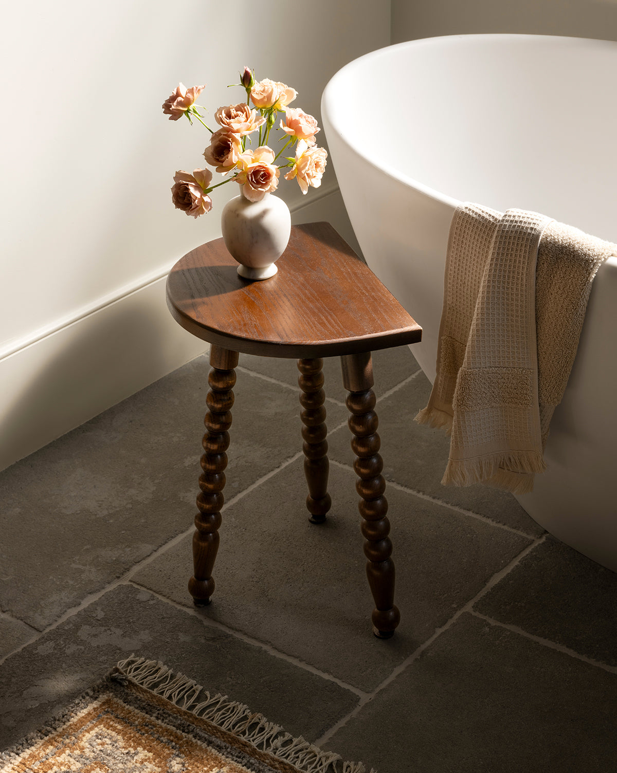 A small wooden side table with three carved legs holds the Fera Bud Vase filled with peach roses beside a white bathtub. A cream towel hangs on the tub, and a woven rug lies on the stone tile floor, by McGee & Co.