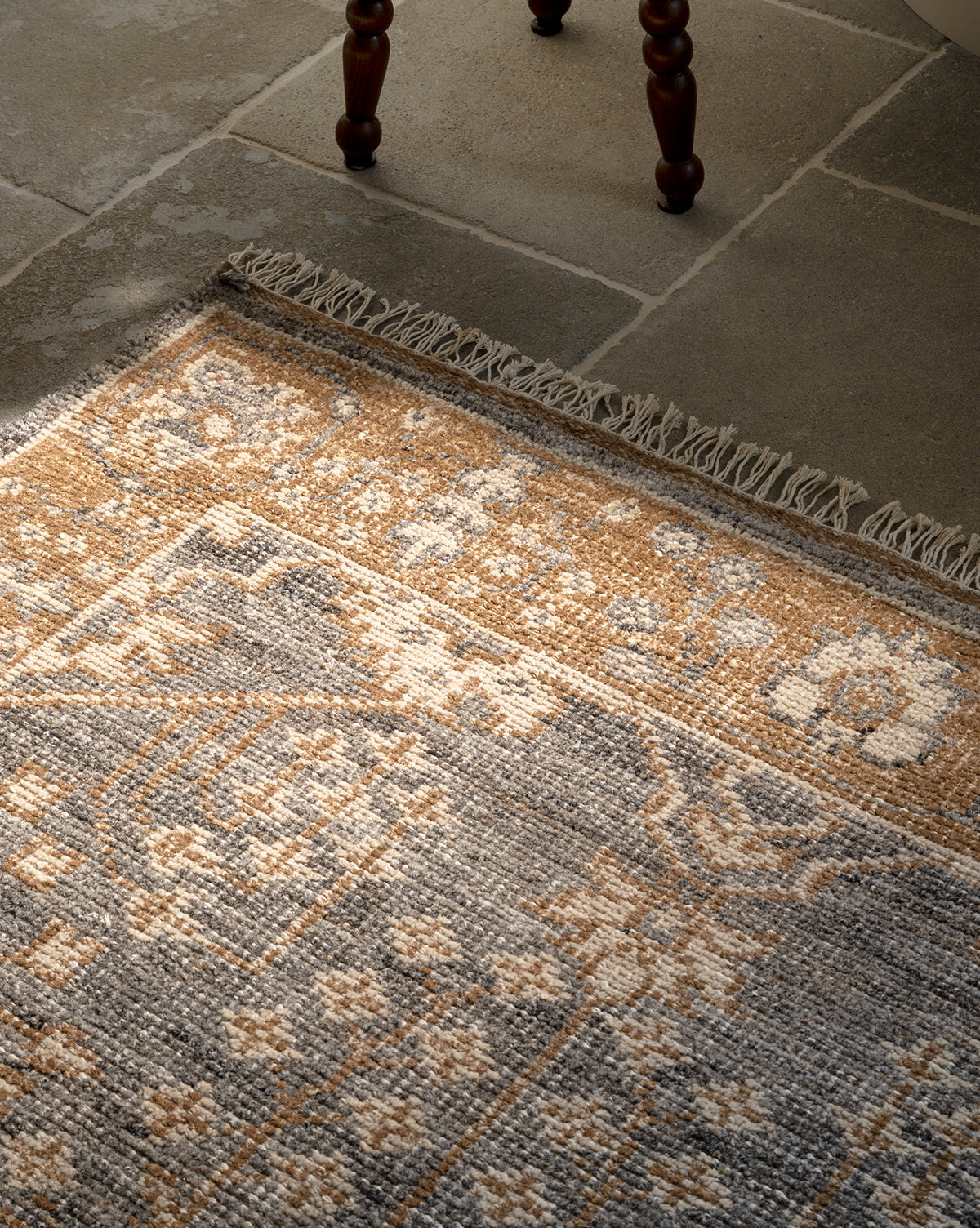 A close-up of the Elison Hand-Knotted Wool Rug in beige, brown, and gray on stone tile flooring, showing artisan details like fringed edges and a wooden chair leg at the top, by McGee & Co.