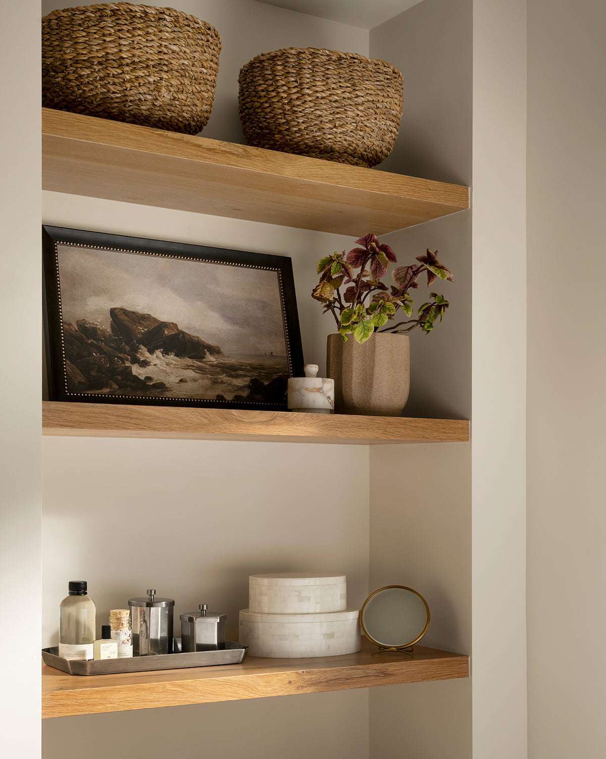 Three wooden shelves display woven baskets, a framed seascape, a potted plant, the Verona Matte Marble Lidded Jar, small bottles, a tray, and a round mirror—perfect inspiration for elegant kitchen countertop decor, by McGee & Co.