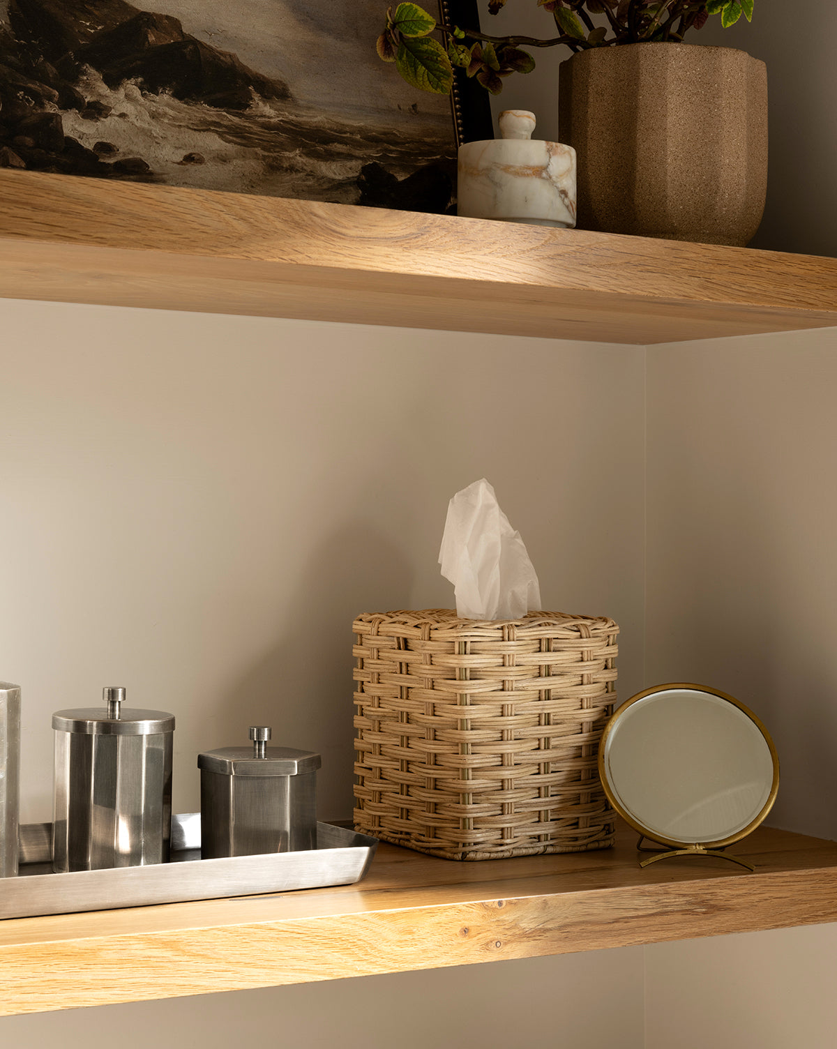A tissue box with a woven basket cover sits on a wooden shelf beside a small round dresser mirror, metal containers on a tray, a plant, artwork, and the Antique Brass Folding Mirror on the upper shelf, by McGee & Co.