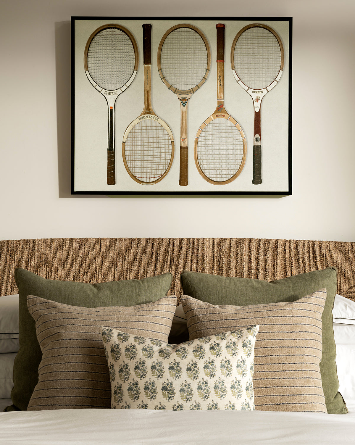 The Rackets 1 giclee print, featuring five vintage wooden tennis rackets, hangs above a neatly made bed with green and striped pillows, a floral cushion, woven headboard, neutral bedding, and a black woodgrain frame, by McGee & Co.